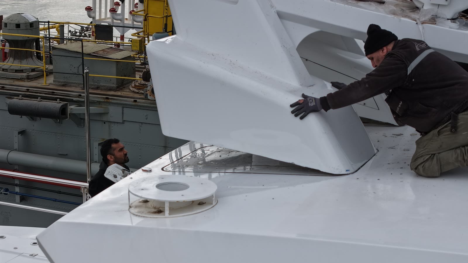 Two fitters in winter clothing lifting a large white moulded foredeck panel back into its recess on a yacht foredeck, a grey commercial vessel alongside behind them