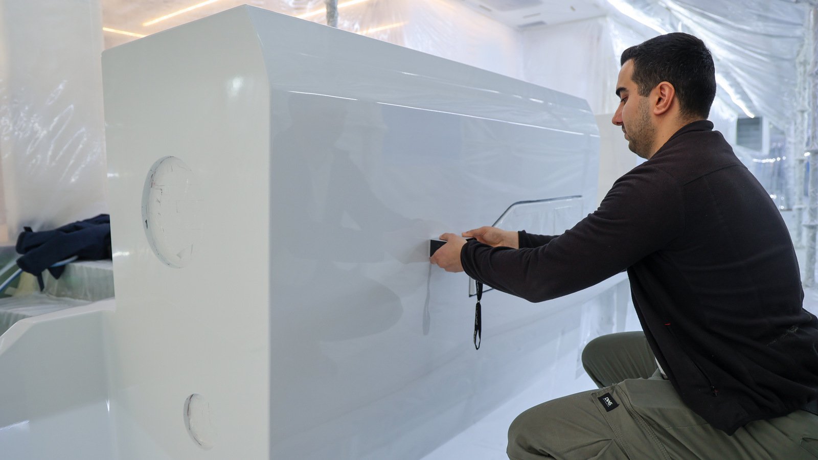 Inside a masked-off paint tent draped in clear polythene sheet with LED strips along the overhead, a man in a dark fleece and olive work trousers kneels beside a large component finished in high-gloss white, holding a small black handheld instrument flat against its side to take a reading, a black wrist lanyard hanging from it; two circular filler patches faired flush show as faint pale discs on the end face.