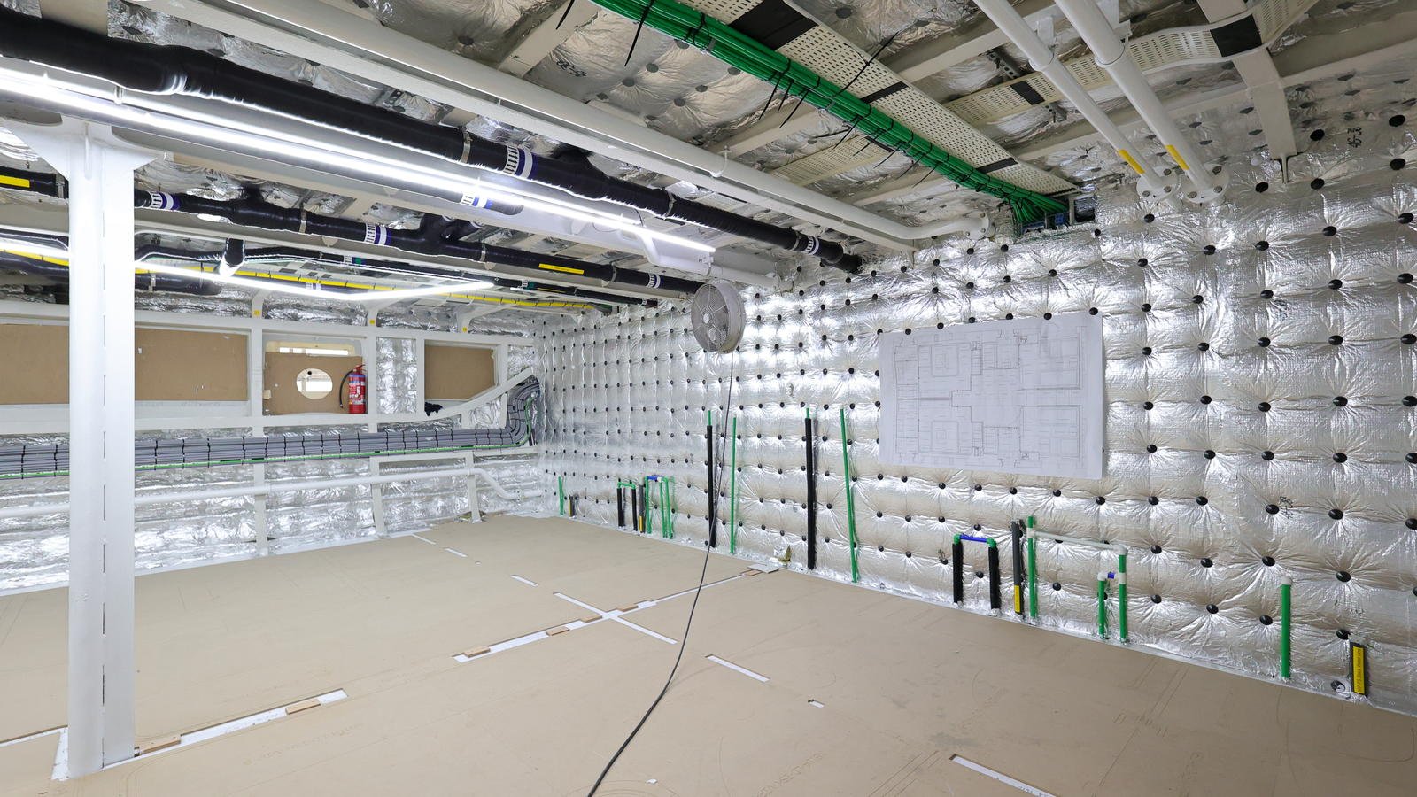 A large interior compartment stripped back during a refit: foil-faced insulation pinned to the shell and deckhead, white-painted deck beams and cable trays overhead, black lagged ventilation trunking and bundles of green cable, white pipes with yellow identification bands crossing the space, and capped pipe stubs in black, green and blue standing proud of the lining along the right-hand side, waiting to be connected.