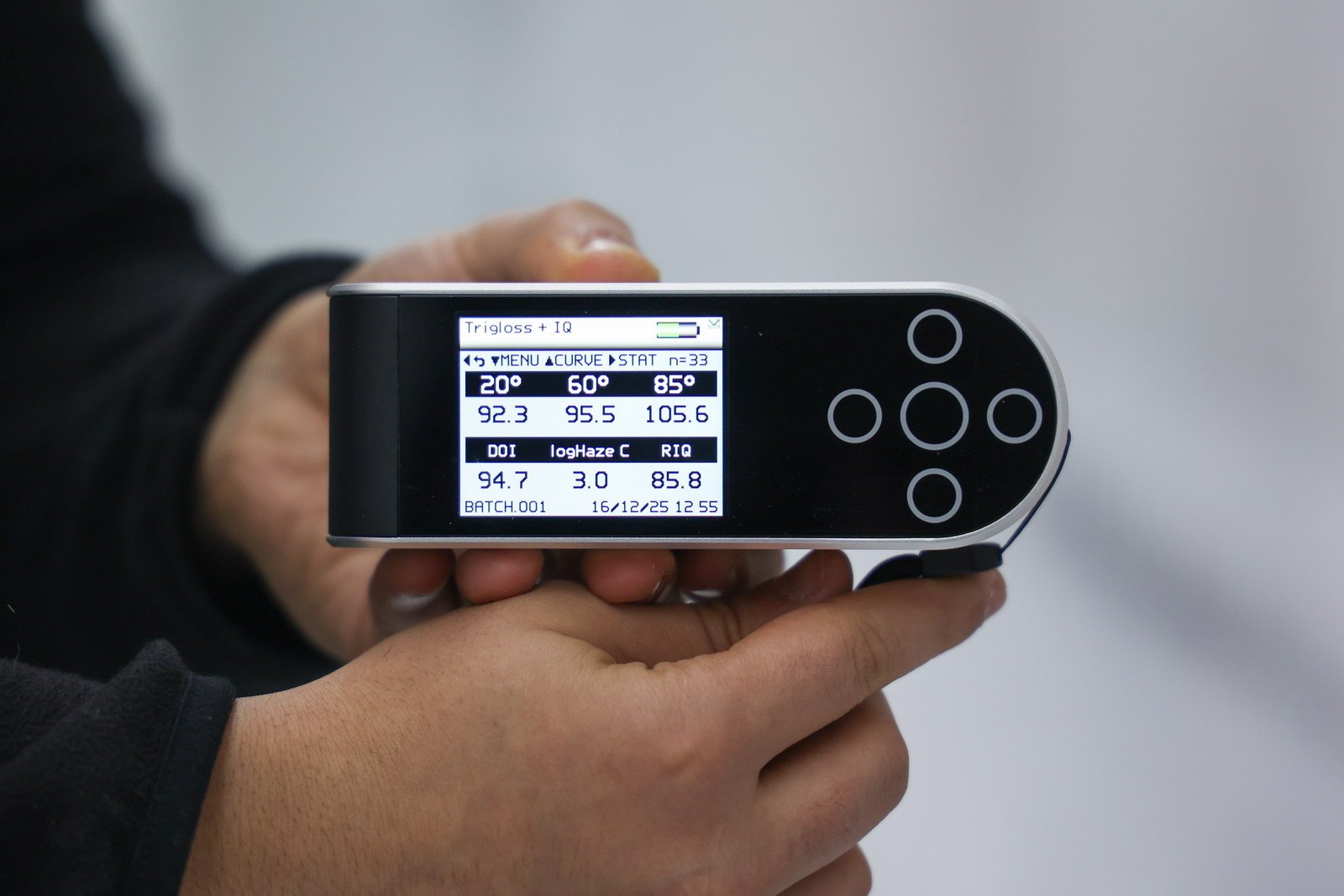 A hand holding a gloss meter against a finished topcoat, its screen showing readings at 20, 60 and 85 degrees