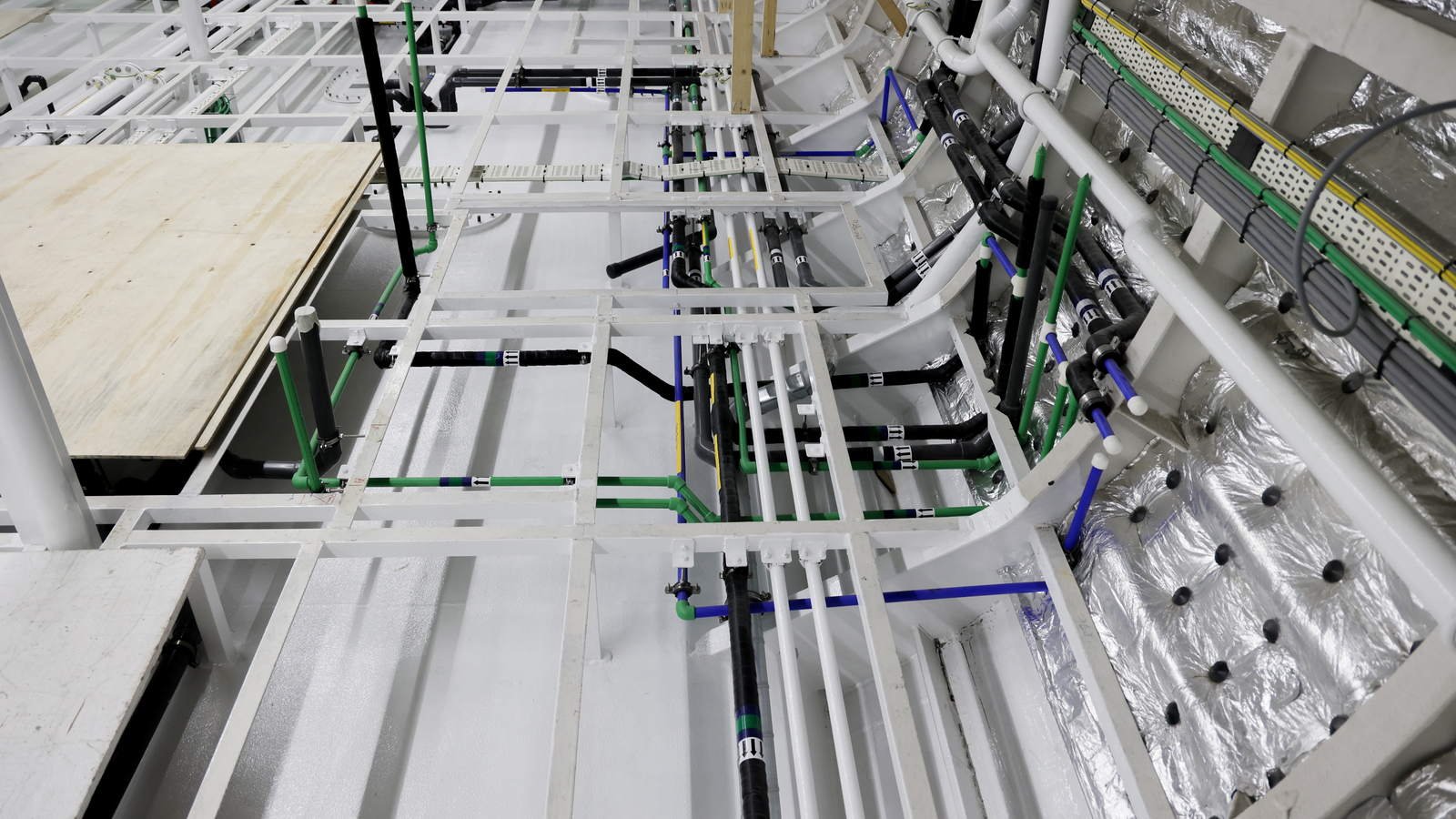 Colour-coded and labelled pipe runs and cable trays carried through a void, with insulated ducting along the shell