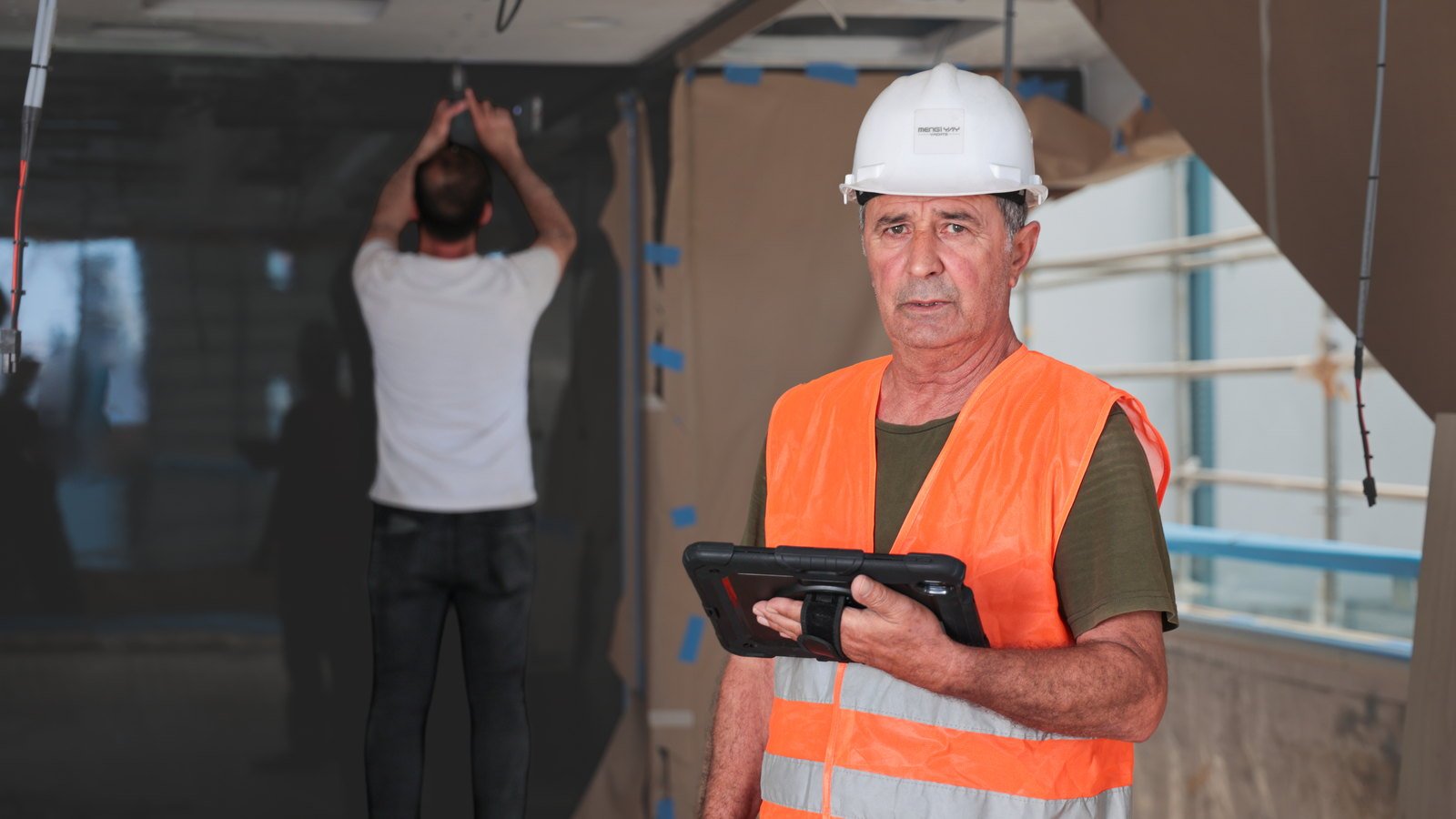 A yard supervisor in a hard hat and high-visibility vest recording work on a tablet inside a stripped interior, a tradesman masking a bulkhead behind him