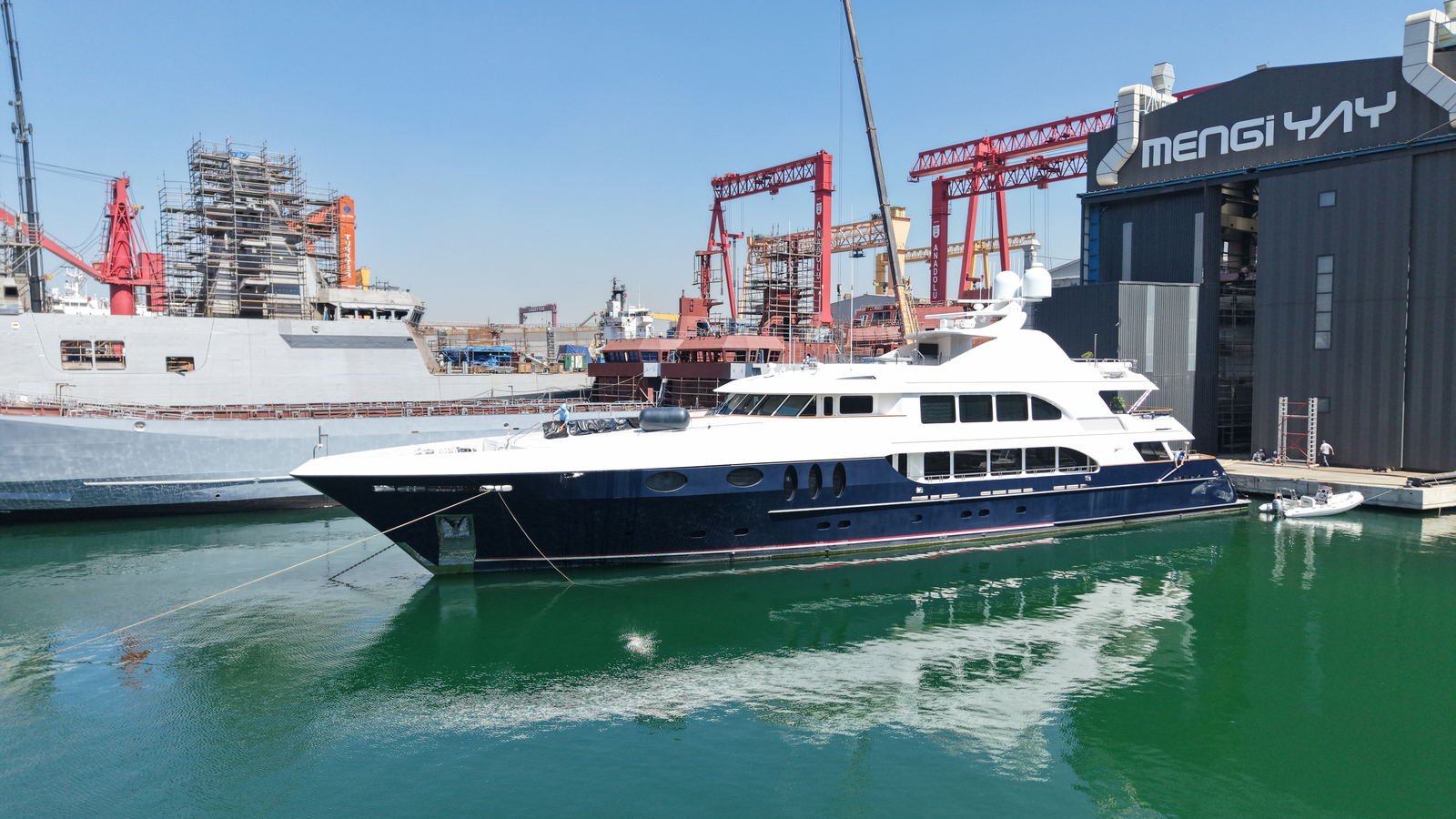 A superyacht alongside the Mengi Yay shed at Tuzla, with commercial shipbuilding gantries and a hull under construction behind her