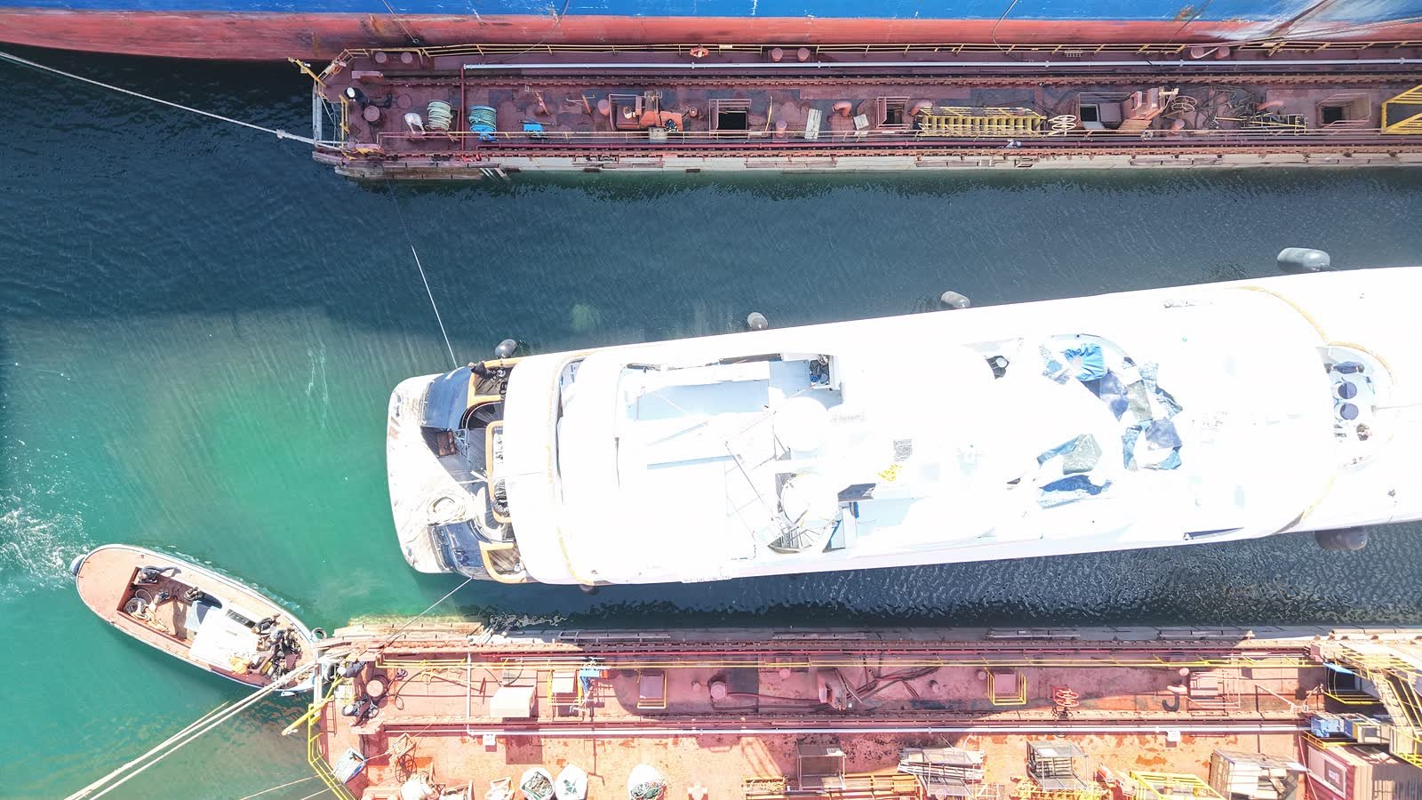 A motor yacht lying alongside a rusted commercial hull in a Tuzla repair berth, seen from directly above, a workboat made fast at her bow