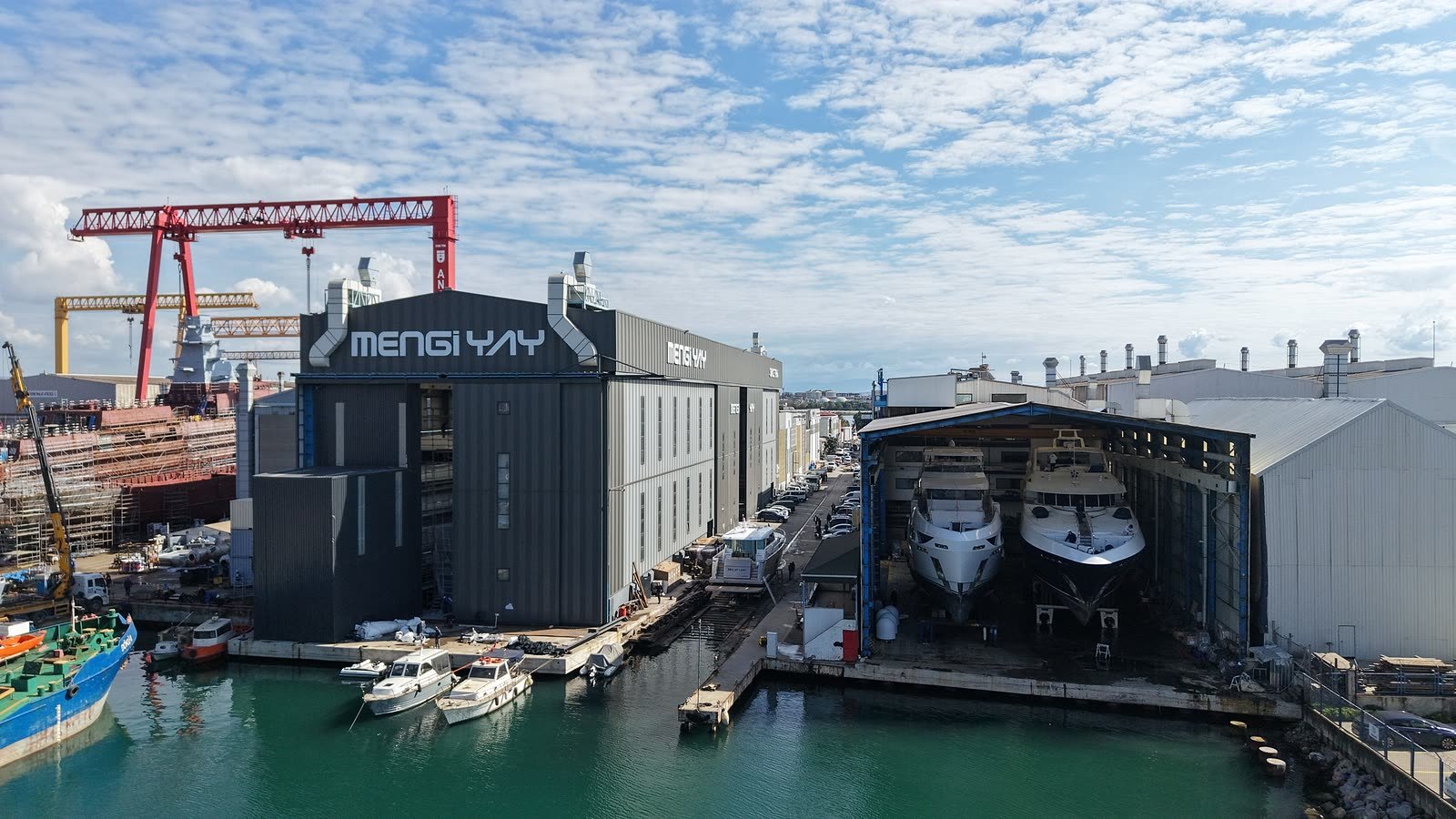 Covered refit halls at Tuzla, two motor yachts inside on cradles with staging up their length, a shipbuilding gantry crane behind the shed and small workboats on the waterfront in front