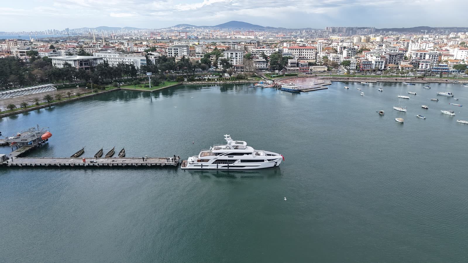 The Tuzla waterfront from the air, a motor yacht lying at a jetty in sheltered Marmara water with small craft on moorings and the town and hills of the Asian shore behind her