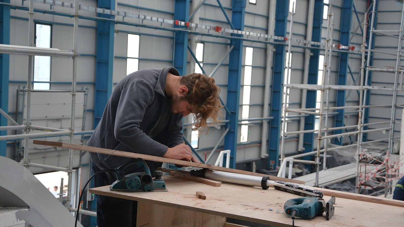 A joiner in a grey fleece leans over a plywood bench inside a refit hall framed in blue steel, marking a thin timber batten with a pencil; a corded electric planer, a jigsaw and a mastic gun lie on the bench beside him, scaffolding towers standing against the wall behind.