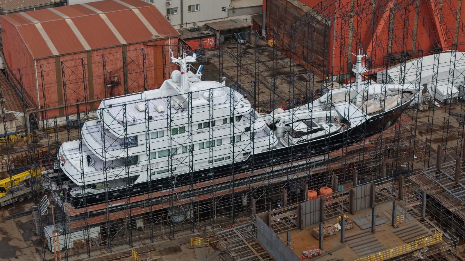 A superyacht ashore inside full scaffolding on the hardstanding at the Tuzla yard, with the red workshop sheds behind her