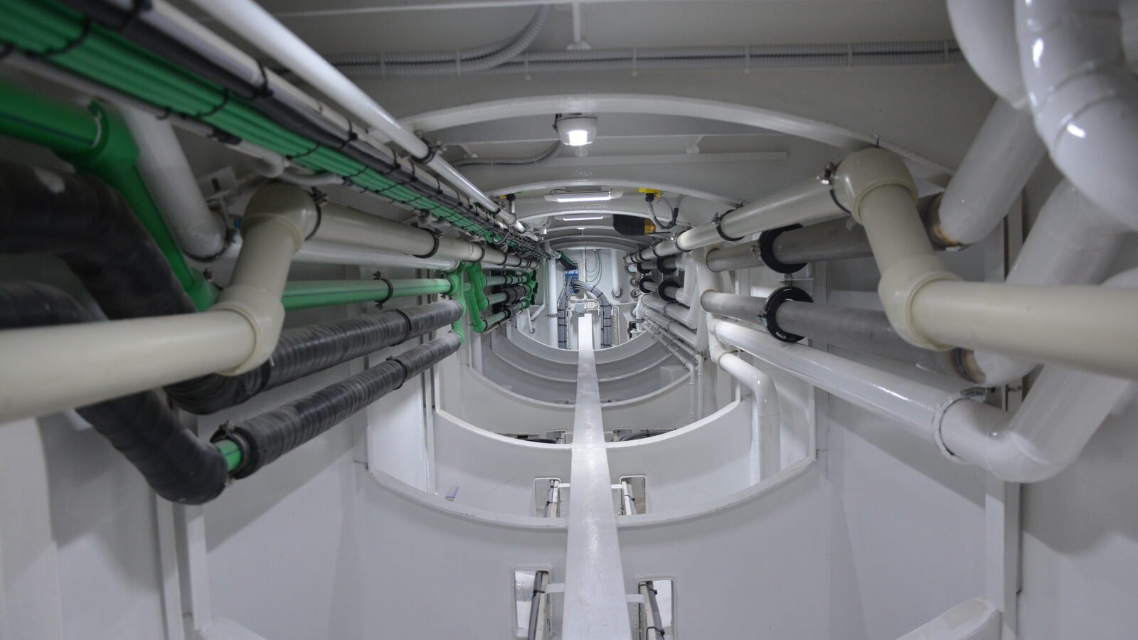 A narrow service tunnel on a yacht's centreline, everything in it painted white, looking fore and aft so that four or five frame rings step away into the distance and close to a small bright opening at the far end. Both sides carry runs of white and cream insulated pipe at about head height, elbowing down and across the frames at intervals and crossing one another where they turn; several lengths are wrapped in black taped lagging, and two or three green plastic lines run high along the left-hand side. A cable tray carrying grey bundles runs along the deckhead on the left, with a flexible conduit crossing the deckhead at the near end. A small round light is fixed to the deckhead close to the camera and a flat strip light further away down the tunnel. A raised white girder runs down the centreline of the floor from the bottom of the frame into the distance, with small brackets set either side of it at each frame. There is no machinery, no ducting and no person in the frame, and no lettering is legible anywhere in it.