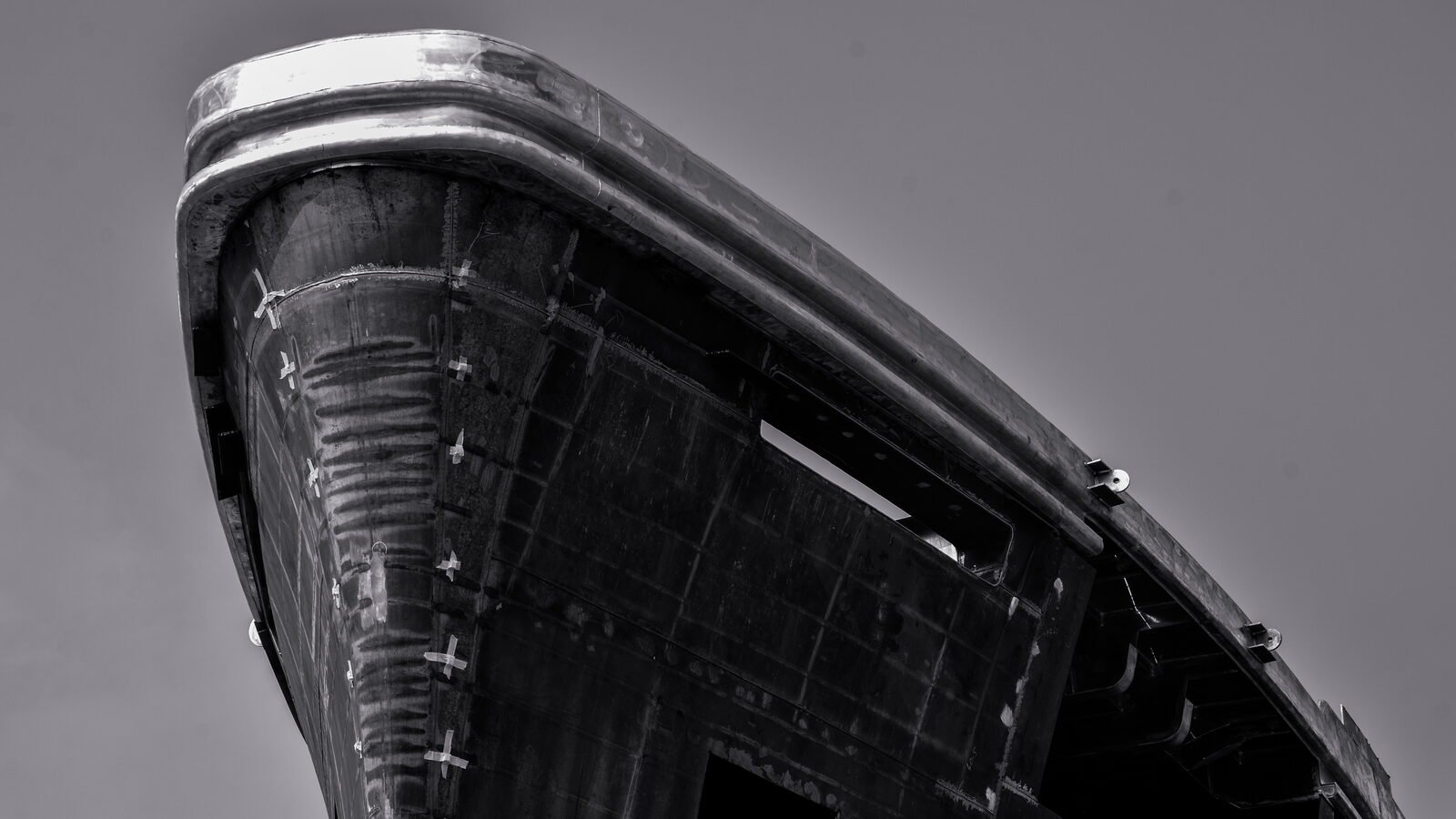 The bare bow of a hull in monochrome, the shell plating showing weld seams and a run of temporary alignment dogs, with an opening cut in the topside plating aft of the stem.