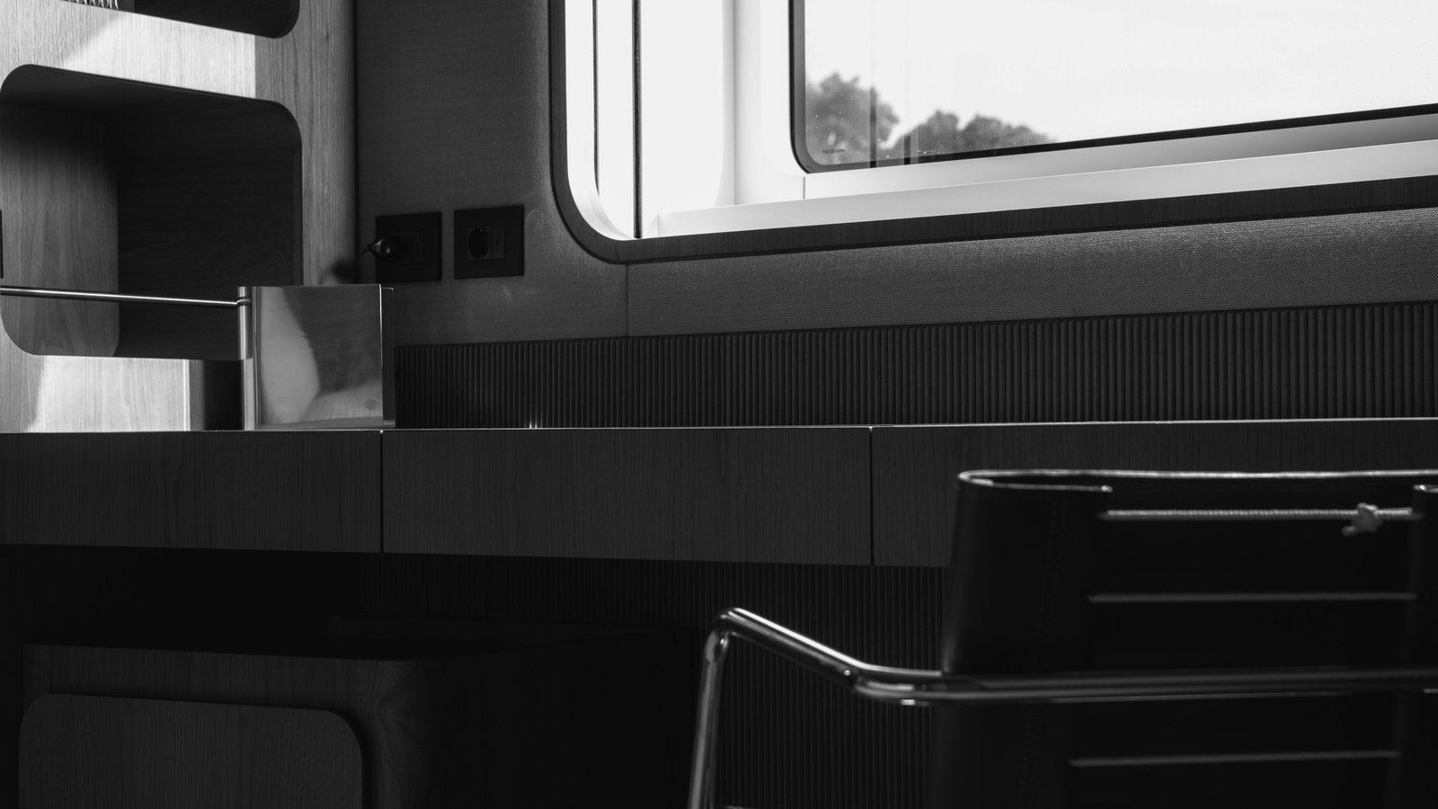 A guest cabin in black and white, the vessel finished and in commission, seen across the whole depth of the desk run: sunlight rakes across an open shelf unit in pale figured timber at the left, a polished metal box stands on the dark timber desk top, and at the right a hull window with rounded corners lets in daylight with out-of-focus foliage beyond. Two flush electrical sockets are set into the timber beside the desk, whose top runs the width of the frame with visible joints between its sections and a drawer unit below, and a black leather cantilever chair on a slim chromed frame stands in the right foreground. A band of fine dark reeding runs along the back of the desk, with a woven-linen panel and fabric surround at the window. Every surface is finished, unmarked and in use.