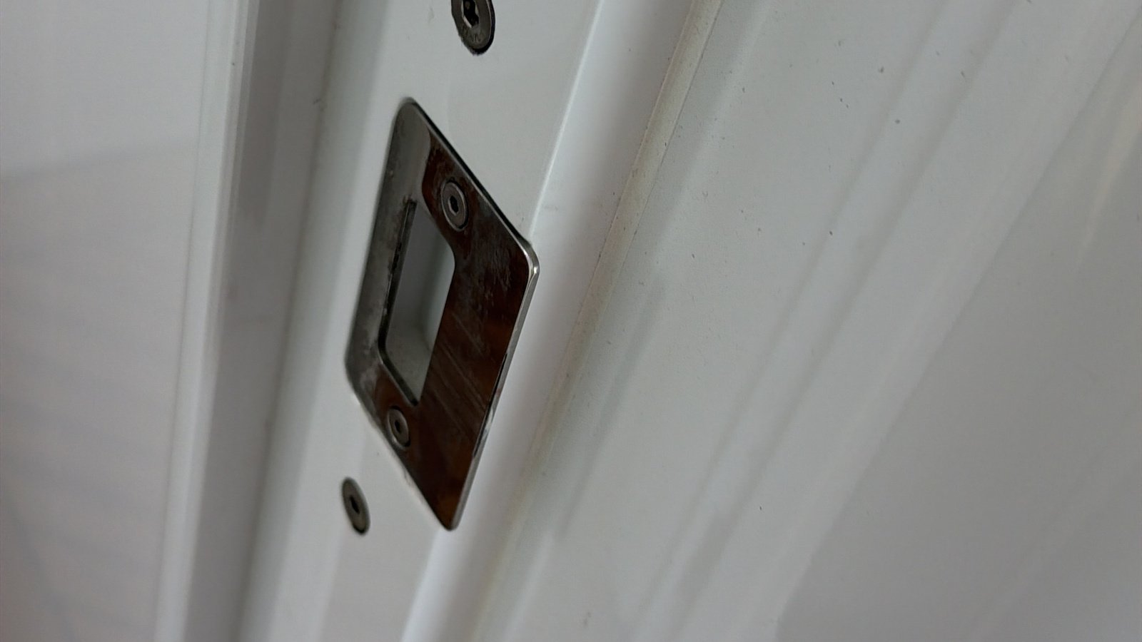 A rectangular chrome-plated striker plate with a slotted keeper, shot upward, set into a white-painted rebate with two crisp parallel beading mouldings running away to the right, the plating broken down to mottled brown and grey-green corrosion bled from its lower edge onto the white paint, two dark corroded countersunk screws in their recesses, a single empty grommeted screw hole to the right and a row of four or five small dark fastener holes further along where a fitting has been removed, the white gloss around it sound but lightly scuffed and varnished timber edges showing at the top right and bottom right corners.