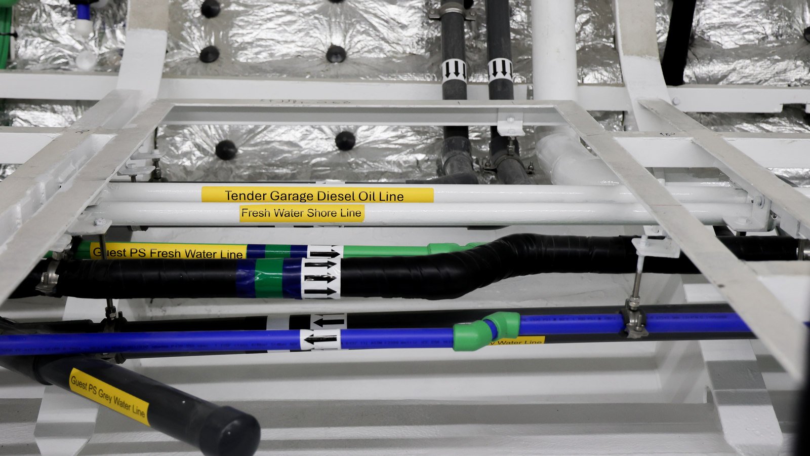 Service runs carried on a white-painted pipe rack against foil-faced insulation, each line named on a yellow adhesive label: "Tender Garage Diesel Oil Line" and "Fresh Water Shore Line" on two white pipes, "Guest PS Fresh Water Line" on a black lagged line and "Guest PS Grey Water Line" on a black pipe running down to the left, with a blue pipe and a green pipe with a fitted coupling crossing the frame, white flow-direction arrow bands on two of the lagged lines, and stainless steel clamps holding everything to the rack.