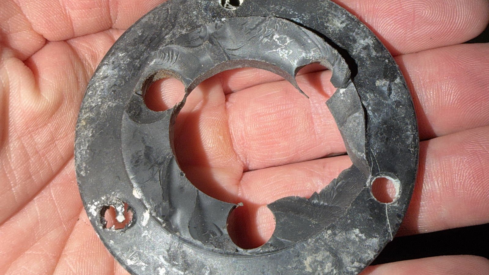A corroded circular backing ring held in an open palm, four fastener holes around its rim, its raised inner collar cracked clean through and missing a section, grey-black corrosion product over most of the surface with white crystalline deposits around the holes, two of them corroded out of round, against a dark workshop background.