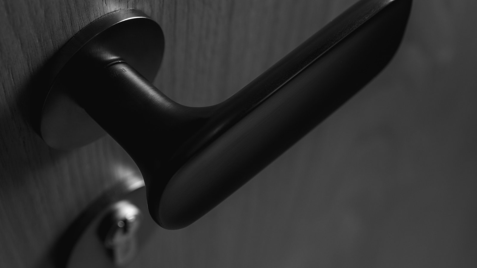 Black-and-white close view of a cabin door: a matt-black lever handle mounted on a round rose against dark timber with a visible open grain, the lever sweeping to the right with a soft highlight along its underside, and below and slightly left, out of focus, a polished metal escutcheon carrying a keyway.