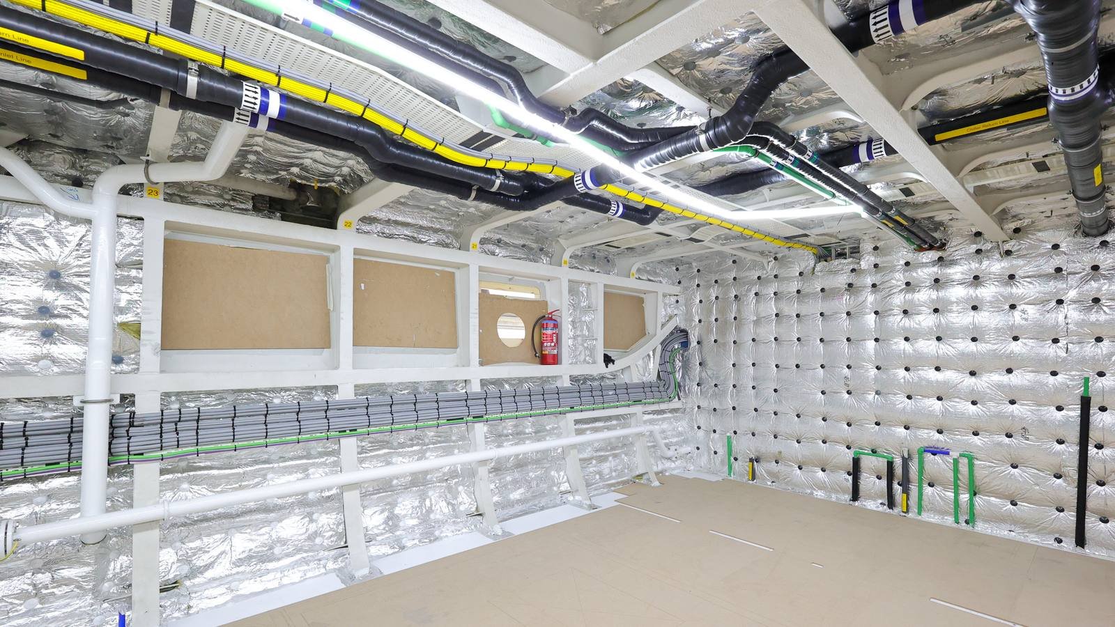 A stripped interior space during fit-out, with bare framing and foil-faced insulation on the deckhead and bulkheads, plywood panels stacked against one side, a bare floor, and a long grey perforated cable ladder running horizontally along the bulkhead just below the deckhead