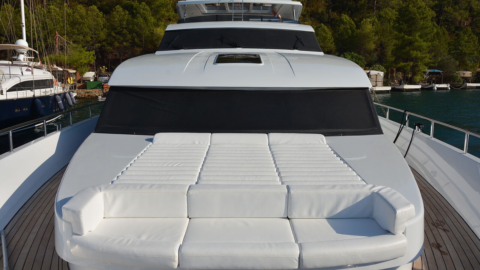 A foredeck seen from the bow looking aft: a curved white upholstered settee with piped seat and back cushions, and behind it a raised sunpad in three channel-stitched sections.