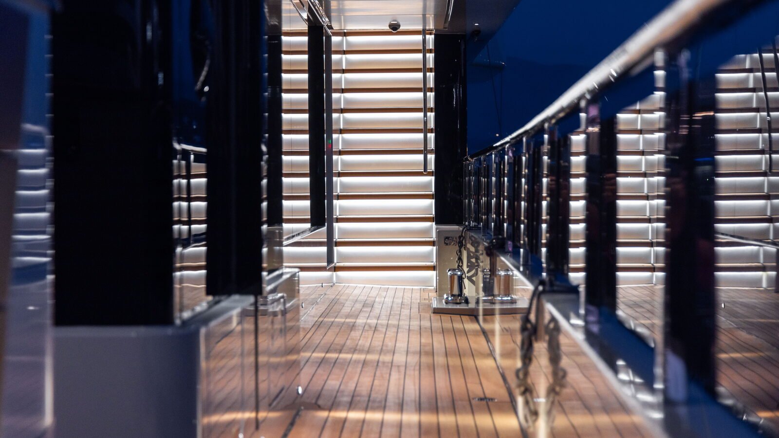 A narrow side deck at night, teak underfoot, a glass and stainless guardrail to seaward and a lit stair filling the end of the passage.