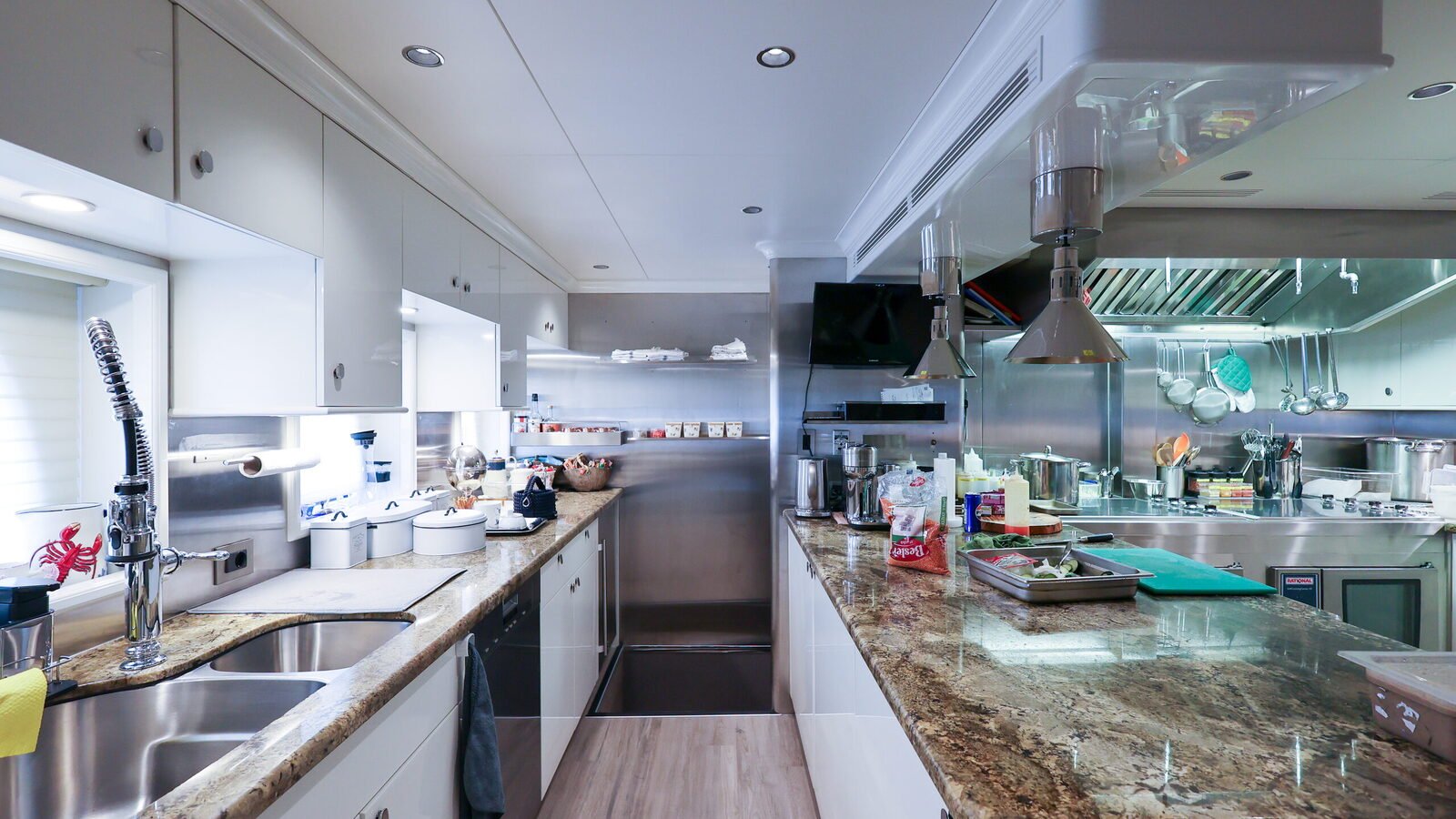 A working yacht galley seen down its length: twin stainless sinks and stone worktops to port, an extraction canopy and heat lamps over the range to starboard, and a ventilation grille in the deckhead.