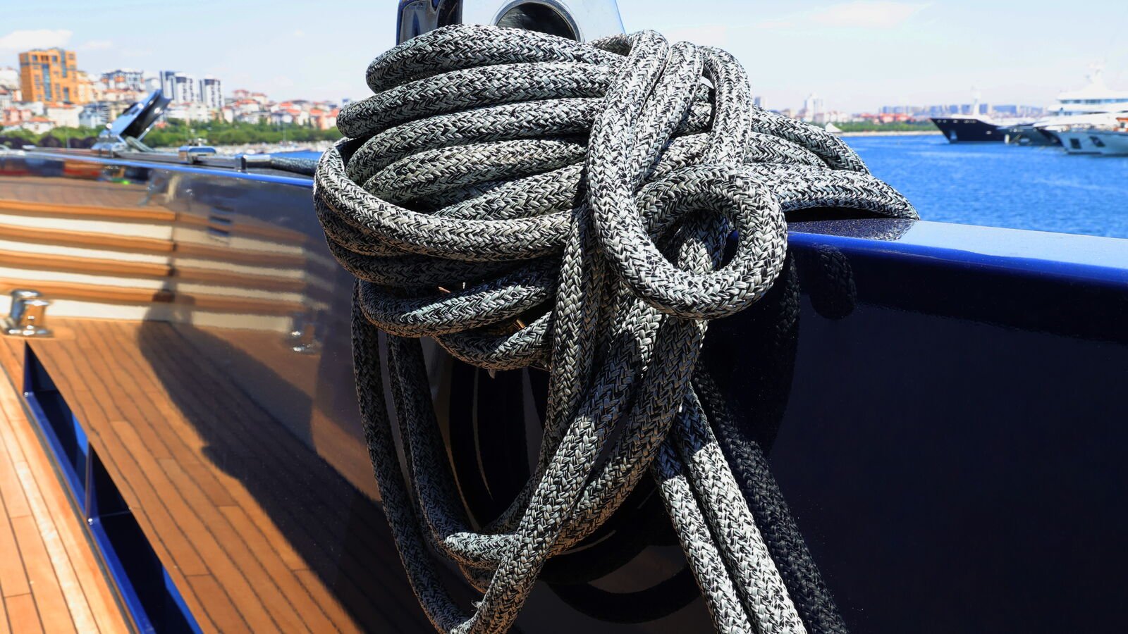 A braided mooring line made up in a figure-of-eight over a polished stainless deck fitting on a varnished teak side deck, a dark blue bulwark falling away beyond it.
