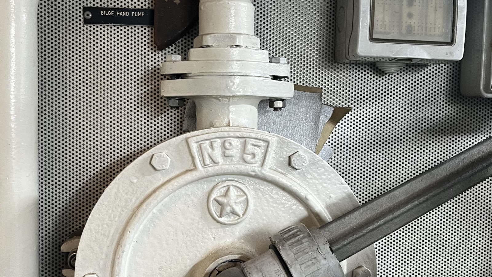 A heavily white-painted cast pump mounted against a flat metal panel punched with a dense regular grid of small round holes, which fills the frame edge to edge behind it. The pump's circular cover plate dominates the lower half, the paint thick enough to have softened every edge and left a hammered texture across it; cast into the cover and painted over with it are a raised rectangular cartouche reading No 5 and, below that, a raised five-pointed star inside a raised circle, a device carrying no wording. A grey steel bar enters from the right-hand edge and runs out of the picture, socketed into a cast collar at the centre of the cover and clamped there by a serrated band. Above the cover a white painted riser descends into a pair of bolted flanges, the nuts and bolt heads painted over with everything else, and a plain painted pipe runs down the extreme left edge. At the upper left, screwed to the perforated panel, is a small black engraved label in white capitals reading BILGE HAND PUMP. At the upper right is part of a grey weatherproof electrical enclosure with IP55 moulded into its case.
