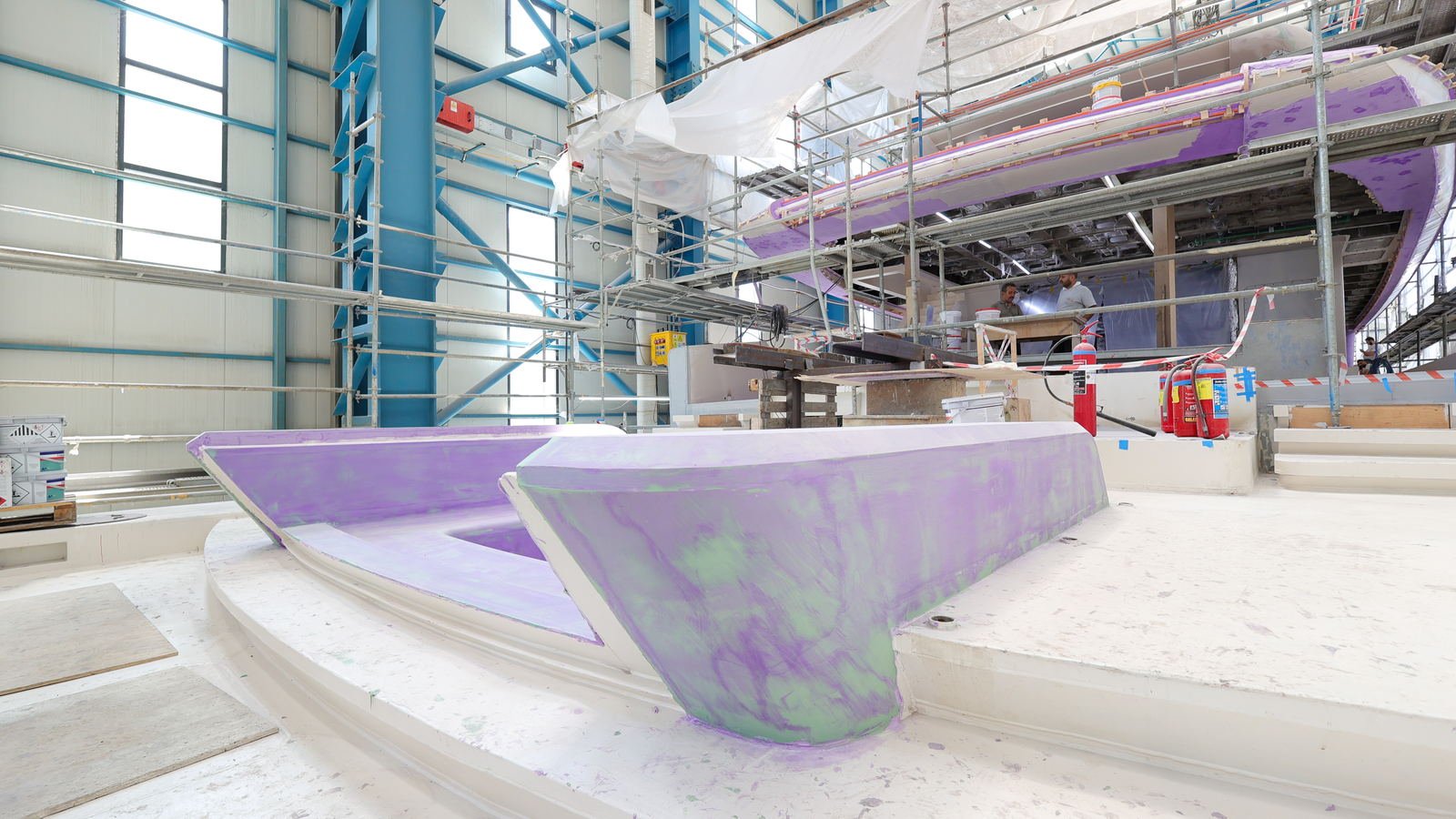 Hull and superstructure sections under scaffolding inside a covered hall, coated in purple and green fairing compound, with two men working at a bench beyond
