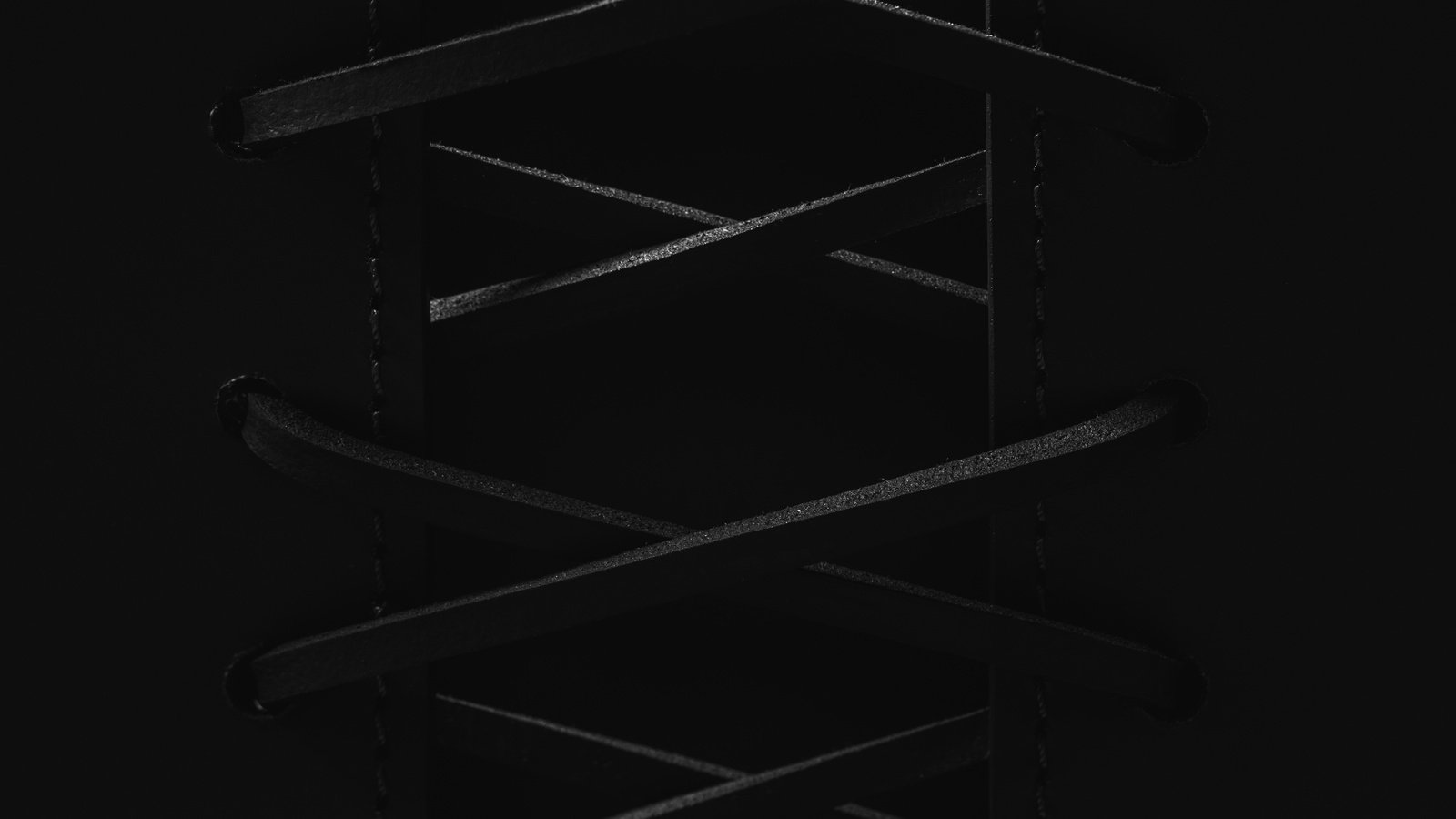 Very low-key monochrome macro, nearly black. Two dark hide panel edges stand apart down the middle of the frame with a broad opening between them, roughly two fifths of the picture width, and the two edges are never brought together. Flat cut thongs turn back on themselves in rounded returns set well back in each panel face, with no metal ring or collar visible at any of them, then cross the faces and lace back and forth across the opening in X crossings, the returns evenly pitched down each panel. Two crossing points are complete down the centre of the frame; a third sits just inside the bottom edge with its limbs running out of frame, and a fourth falls above the top edge, where the two thongs leave the frame before they meet. The brightest parts of the picture are the lit top faces of the thongs and the fine lines along their long upper edges, running the width of the frame, while the rounded returns catch far less. A fine dotted saddle-stitch line runs vertically down each panel, between its column of returns and the opening, and beyond each column the hide runs on unbroken and matte to the sides of the frame, at the same near-black as the rest.