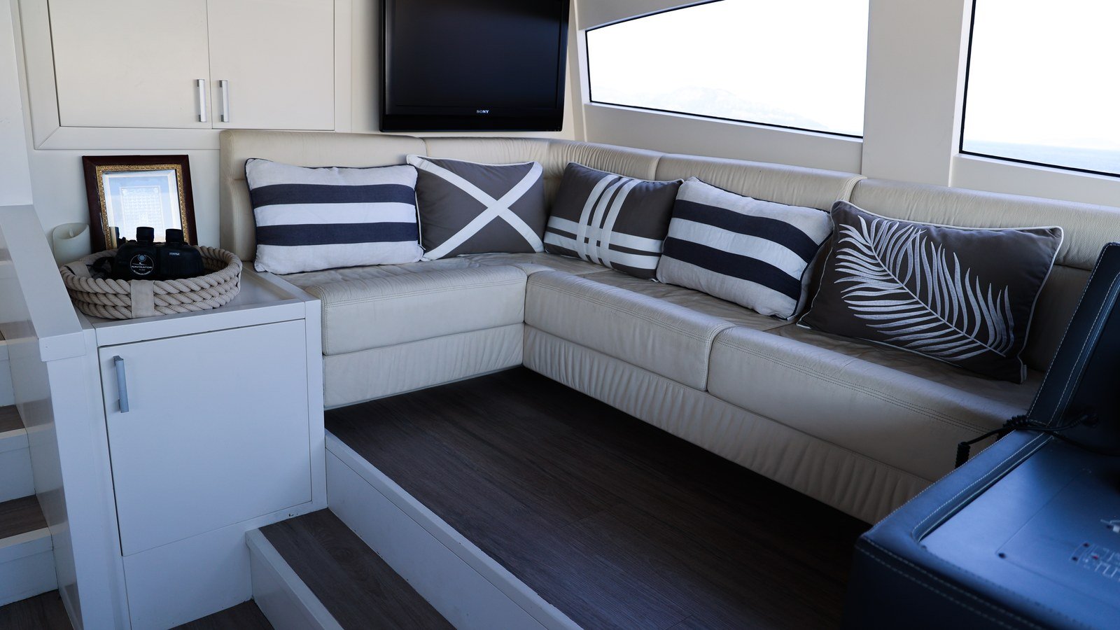 A yacht saloon settee in pale leather against fitted joinery lockers and a timber sole, with scatter cushions and a long side window