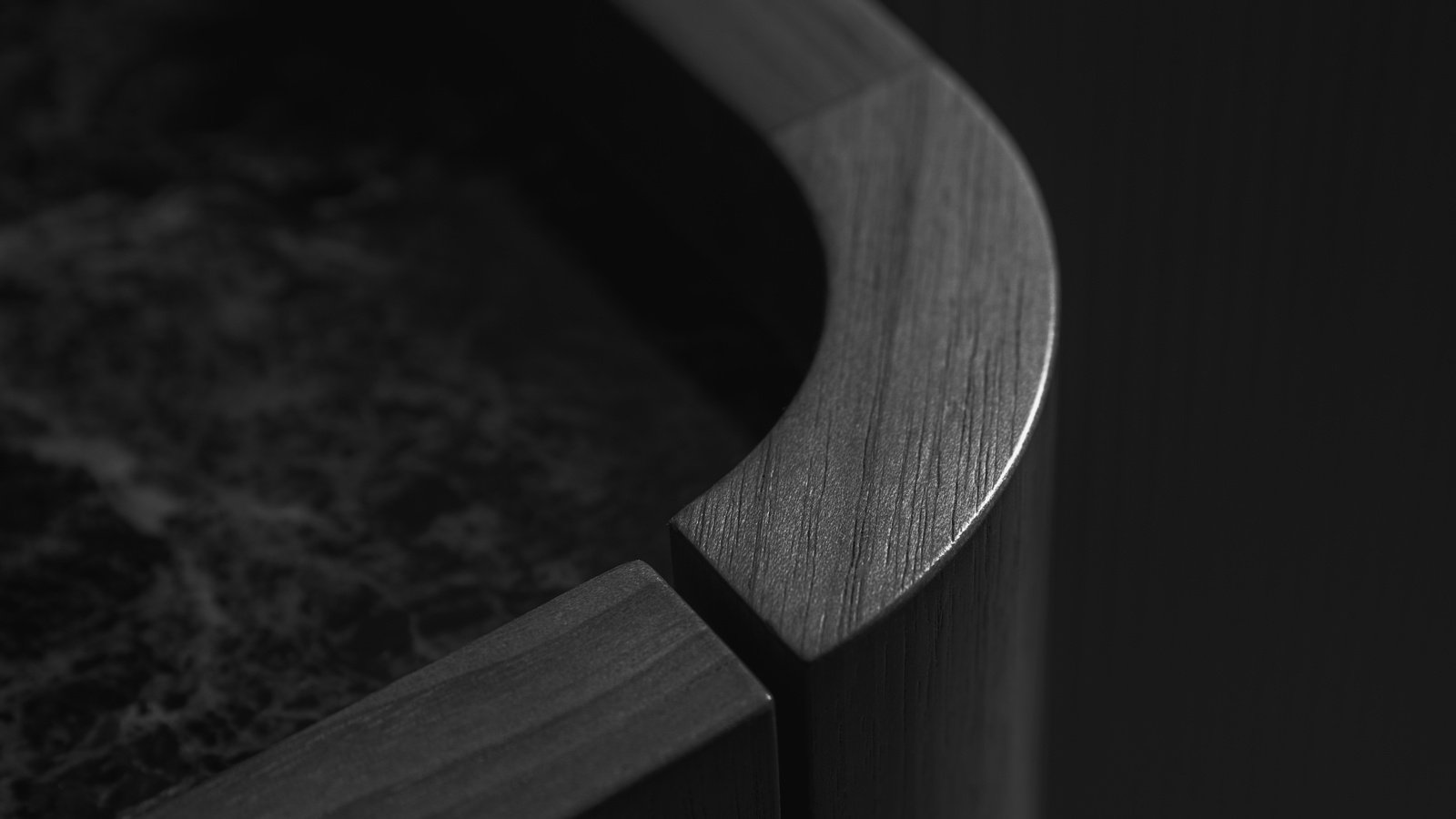Monochrome close view of a joinery corner, lit from the right so the outward-facing surfaces of the sweep are the brightest things in the frame. A solid timber rail with a flat top face and eased arrises down each side curves round the corner from the lower centre out to the right and away off the top of the frame. Its open grain does not read the same way on every face: over the lower part of the sweep the fine streaks run across the width of the top face, while further round, past the joint line, and on the straight length at the lower left, they follow the length of the rail, and on the outboard face below they run down it. One fine dark joint line crosses the rail at the upper right, where the shaped length meets a straight one. At the lower centre there is no joint at all: two square-cut rail ends stand apart at slightly different levels with an open dark gap between them, and the square-cut end faces turned into that gap read as flat dark planes with no grain legible on them. The rail stands proud as a fiddle above the top it retains, a dark surface with pale branching veining, out of focus, running down the whole left side of the frame from near the top left corner to the bottom left. Below and outboard the carcase face falls away smooth and dark as it curves round the corner. The lit face of the rail holds the frame almost to the right-hand quarter, and only beyond it does a flat dark panel begin, filling that quarter as one even field with nothing legible on it.