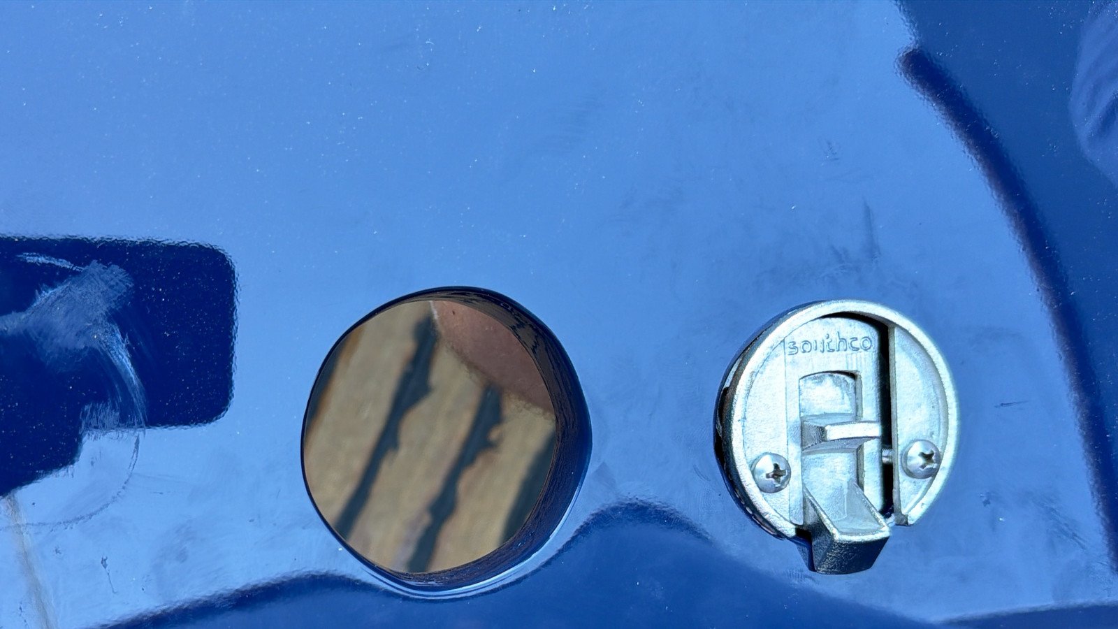 A round aperture cut in a dark blue lacquered panel and set flush in it a polished circular cap, with a flush compression catch let into the panel beside it, its cam tongue set between two crosshead screws. The panel is already in its finished topcoat, the openings cut and the fixings drilled to template afterwards, and hard outdoor sunlight puts pale timber and its shadow lines into the mirrored cap and faint banded reflections across the lacquer, placing the frame in the yard and not in a finished interior.