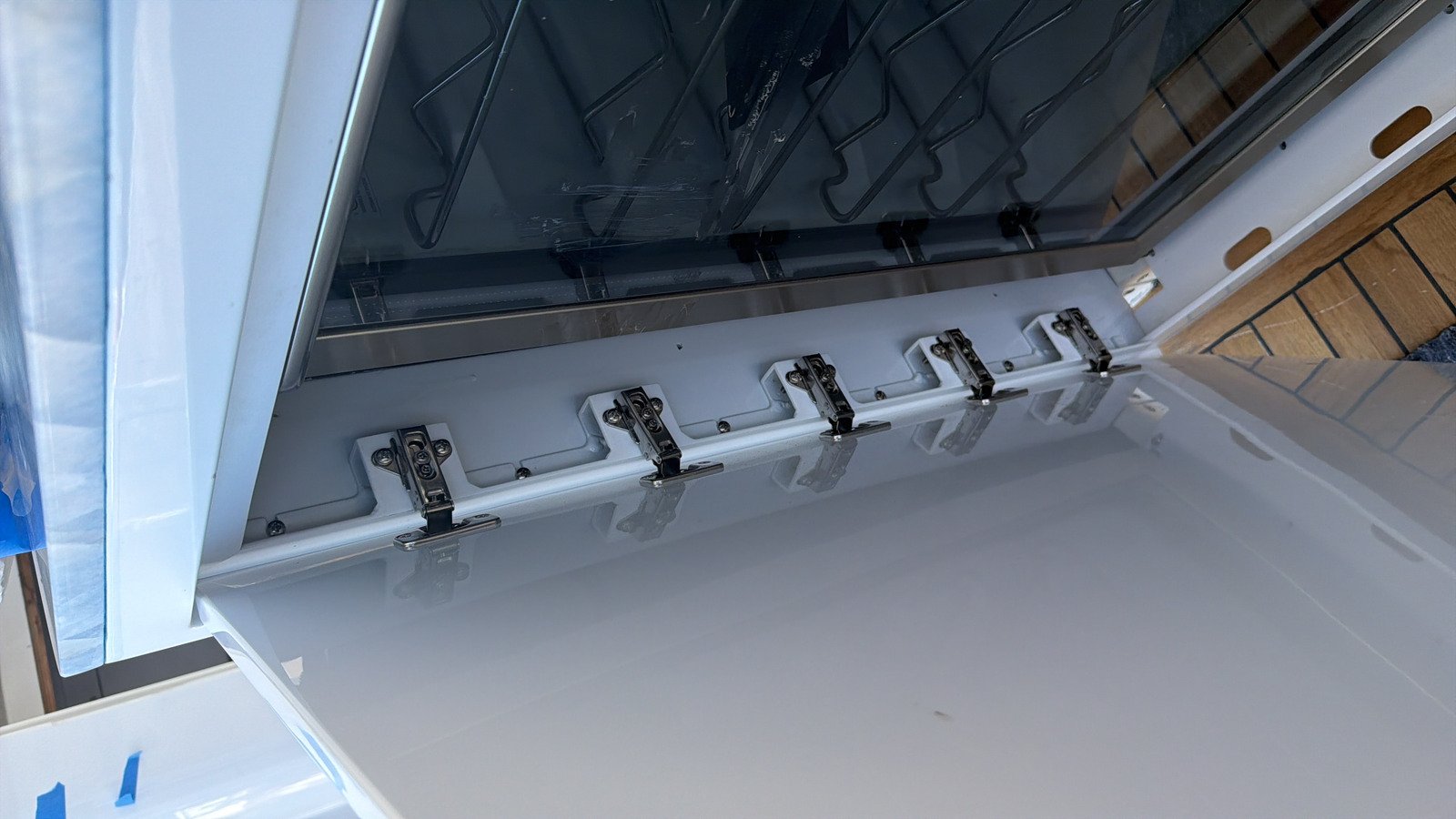A white gloss door standing open in hard daylight, five bright-plated concealed cabinet hinges set in a line along its inner edge, each cup-and-arm body carried on its own mounting plate screwed to a moulded white plinth with its adjustment screws visible on the arm. Beyond the hinge line a metal-framed tinted glass inner door shows dark wire racks behind it, pale blue protective film and sheeting are still taped in place at one edge, and the hinge line is reflected in the gloss white face.