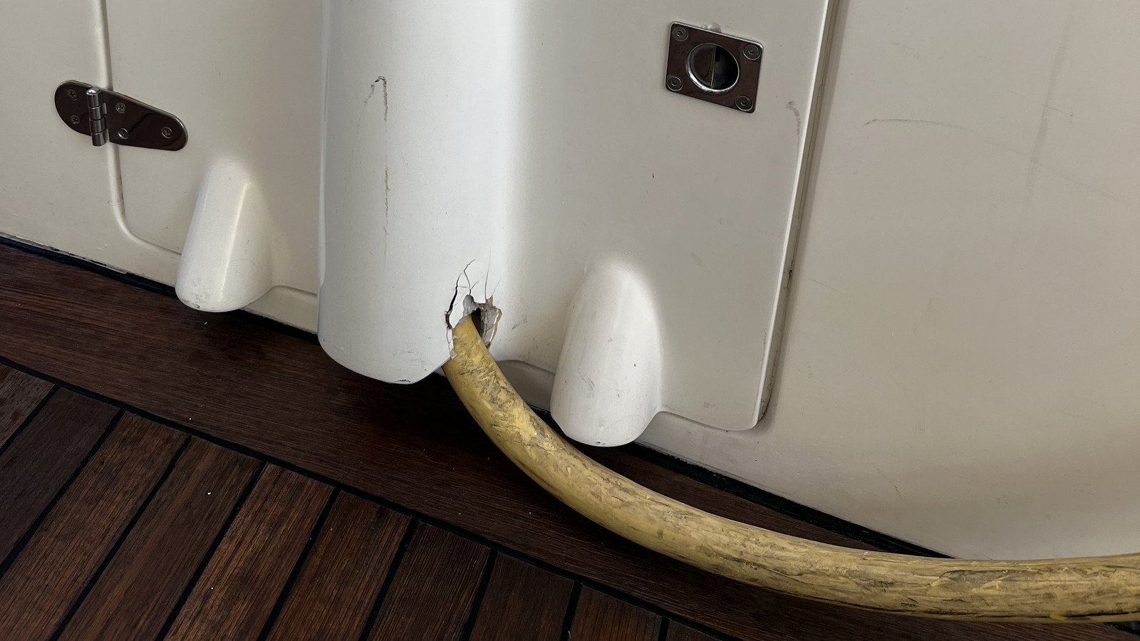 A ragged, torn hole broken through the foot of a white moulded panel, fine cracks radiating from its edges and grey laminate showing where the gloss surface has lifted, with a thick-walled yellow-ochre hose emerging from it. The hose is heavily scuffed and dirty along its length and curves away across the deck to the right, out of the frame. The panel is the lower part of a white gloss moulded unit standing clear of the deck on rounded conical moulded feet, three of them visible, the dark deck showing beneath and between them and a black caulked joint running along their foot. Left of centre a strap hinge spans a vertical joint between two faces of the moulding, its two teardrop leaves dark and patinated over a bright three-knuckle barrel, five crosshead screws visible. At the upper right a polished flush ring pull lies in a rectangular escutcheon with a screw at each corner. The moulding carries grey scuff marks and faint scratches, and the deck below is laid in narrow planks with black seams running diagonally across the lower part of the frame.