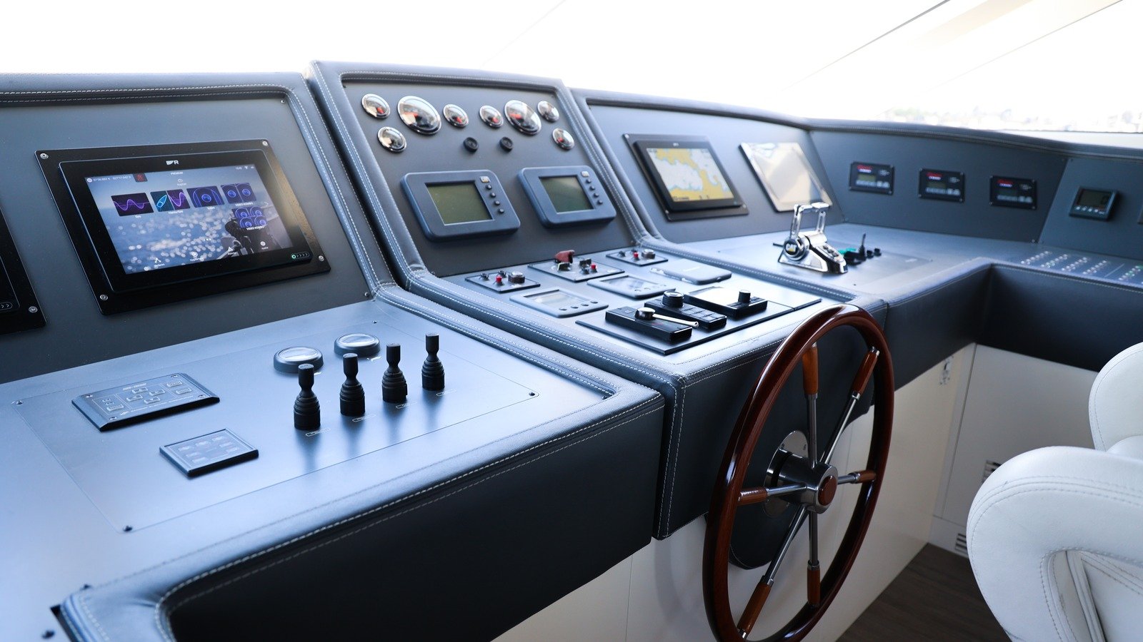 The main-deck console of a motor yacht seen obliquely from port: a chart display and multifunction screens set into a padded dark-grey fascia, a bank of round gauges and switch panels, engine and thruster controls on the sill, a varnished timber wheel and a white helm seat, with daylight through the windscreen