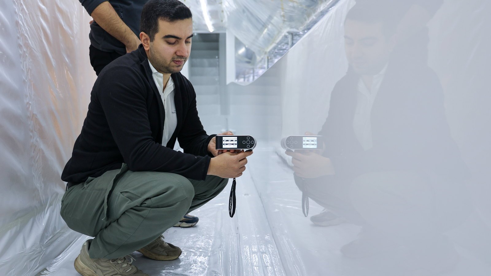 A coatings technician crouched against a finished topcoat inside a taped containment tent, taking a reading with a gloss meter, his reflection visible in the paint