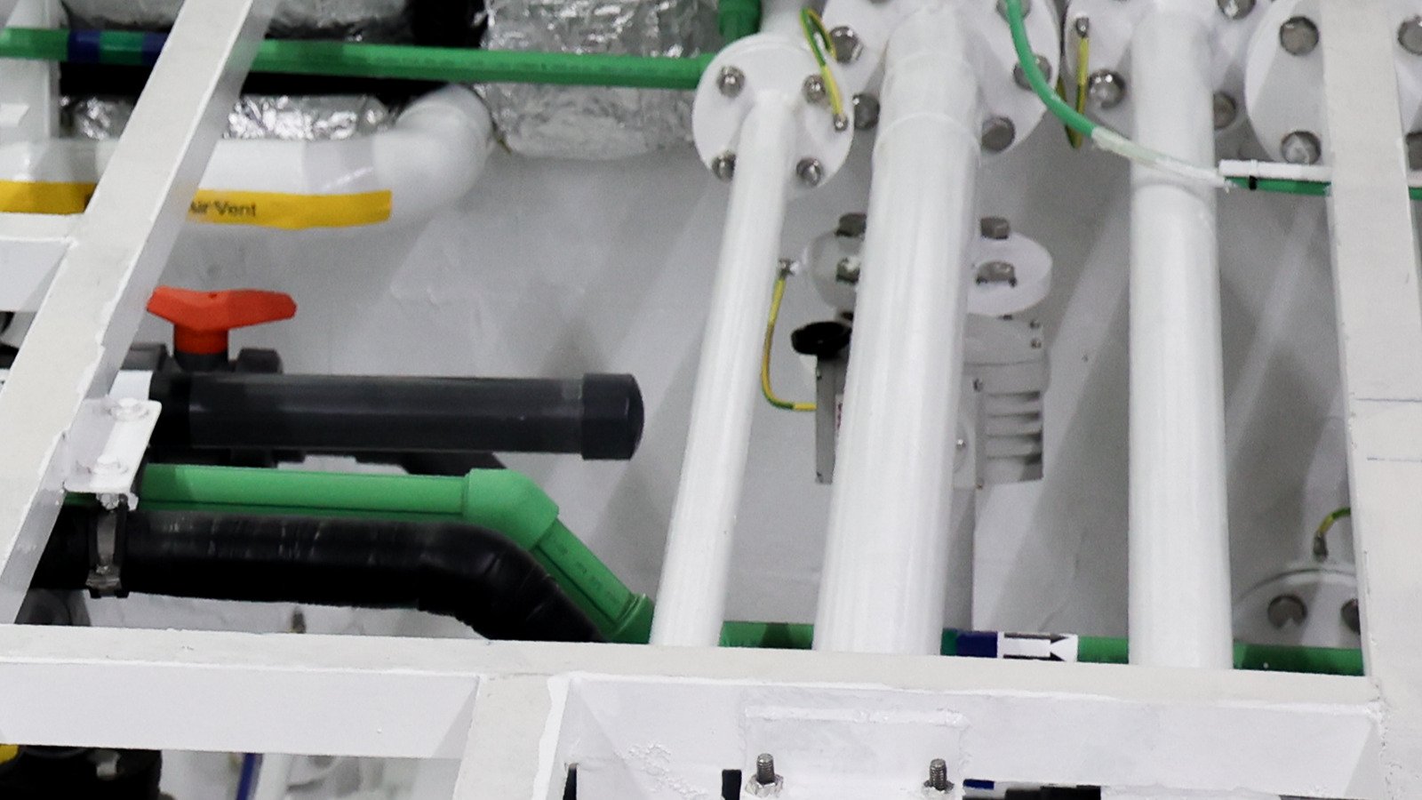 A bank of bolted pipe flanges on a support frame in a technical space, seen square-on at frame level. Four white-painted pipes rise vertically through a white support frame, each made up at a bolted flange with hex bolts and nuts, and three of those flanges carry a green-and-yellow earth bonding strap looped across the joint. At the left a grey ball valve with a red lever stands beside a black capped stub, and above the valve a green plastic pipe turns through a swept elbow over a black lagged line. A small grey instrument body sits between the risers, with white flat-bar supports crossing the frame.