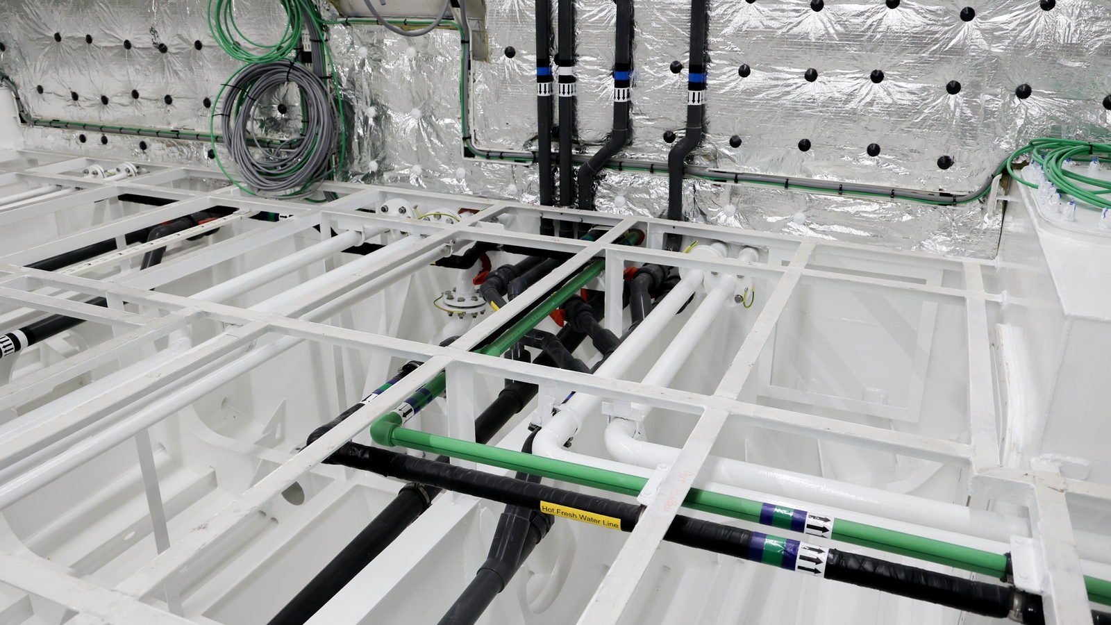 A wide, lower view across a white-painted compartment. In the centre, under a white flat-bar grid, a cluster of grey plastic waste lines runs through swept elbows and clamped couplings; among them a white bolted flange carries a yellow-green earth bonding strap, with two red valve levers behind it and white pipes turning through long sweeping bends, all assembled in the bay. To either side, empty white bays of the structure with nothing in them. Across the top a bulkhead, behind which three black lagged pipes drop vertically with a coiled grey multicore cable and green cable tails hanging beside them. Lower down, green pipe runs cross the frame.