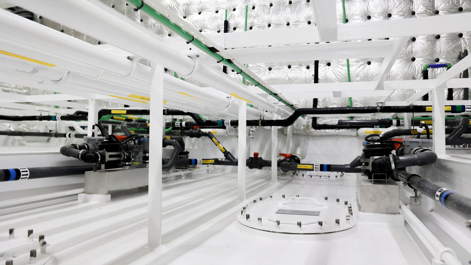 A wide low view across a freshly painted white tank-top space during fit-out. A circular access cover lies flush in the deck at the centre of the frame, ringed with short studs and carrying a small plate, with a second ring of studs at the lower left where another cover sits. Pump-and-valve units stand bolted to low plinths either side, joined to their pipework by flexible couplings and clamps, and black and green runs with identification bands cross the deckhead above against foil-faced insulation.