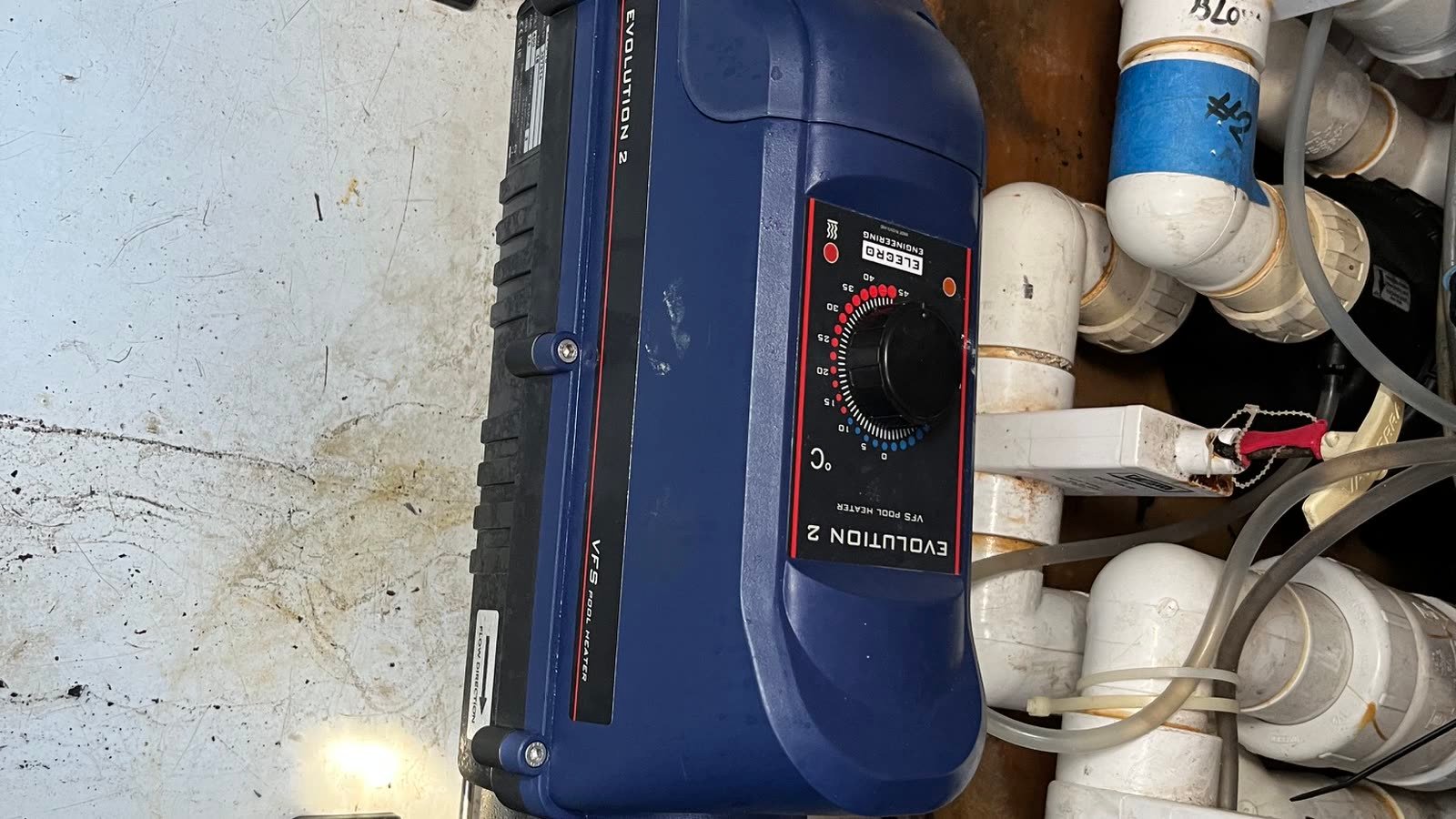 An as-found record photograph looking down into a technical space. A dark blue moulded electric in-line heater lies across the middle of the frame, clamped at each end to a short stainless pressure tube by black bracket clamps, with a black control face and a plain rotary knob on its upper side. To the right a dense bank of white PVC pipework crosses in several directions in elbows, tees and unions, with grey flexible tube, white cable trunking, a small red-handled clamp and a yellow and green earth lead among it. One white pipe carries a hand-written marking and a band of blue tape. The surfaces around it are scuffed white with rust staining at the top of the frame, and a small light shows low on the left.