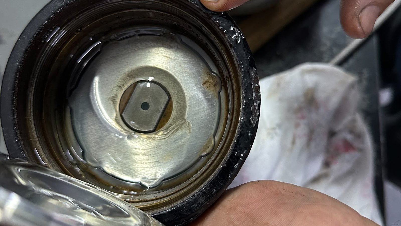 Looking down into an opened cylindrical housing, held up over a bench in ungloved hands, with a fingertip pressed against the rim at the top centre of the frame. Dark oil still stands in the bottom of the housing, and sitting in that oil is a polished machined disc with a dark-edged hexagonal form at its centre and a small round hole at the middle of that, its face showing concentric turning marks and brown staining. The outer rim of the housing is dark and wet, with pale smeared deposits along it. At the lower left a bright, glossy curved form intrudes, too far out of focus to make out. At the right, a single fingertip enters the top corner and rests against a thin bright rod that runs diagonally across it, and below, the side of another hand crosses the lower right, its creases carrying dark specks. A stained white rag and a dark bench top lie soft behind them. One object has become two.