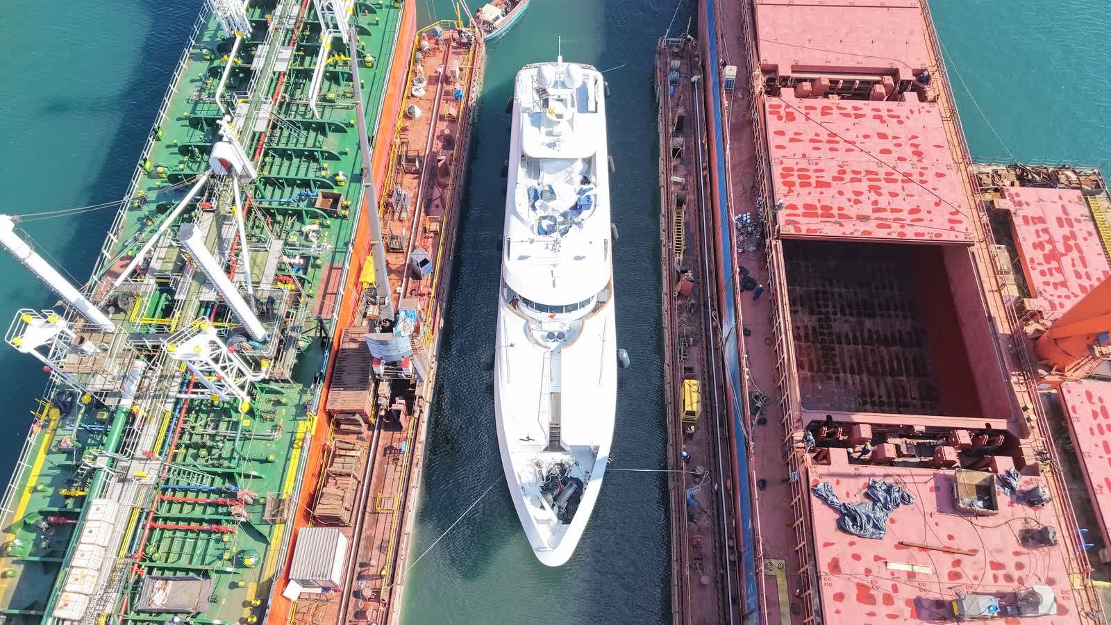 Aerial view of a white superyacht berthed in a narrow channel between two commercial vessels under repair, deck machinery and hatches open on both