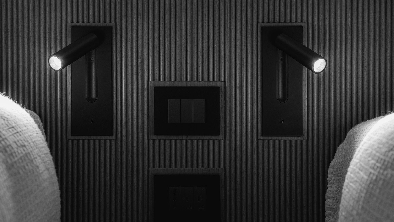 Black-and-white close view of a yacht cabin headboard in finely fluted dark timber, with two matt-black reading lights on swivel arms switched on in flush timber pockets either side and a three-gang rocker switch plate recessed between them, white textured knitted pillows at the lower corners.