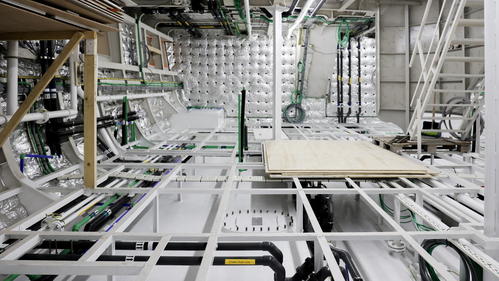 A large white-painted compartment photographed head-on down its centreline, part-way through fit-out: welded white steel framing exposed at sole level and running away from the camera, a stack of plywood sheets laid across it as a temporary working platform, a timber-and-ply staging platform at the top left, a white ladder standing at the right, coils of green cable on the far bulkhead and quilted silver-faced lining on the bulkheads, nothing yet boxed in and nobody in the frame.