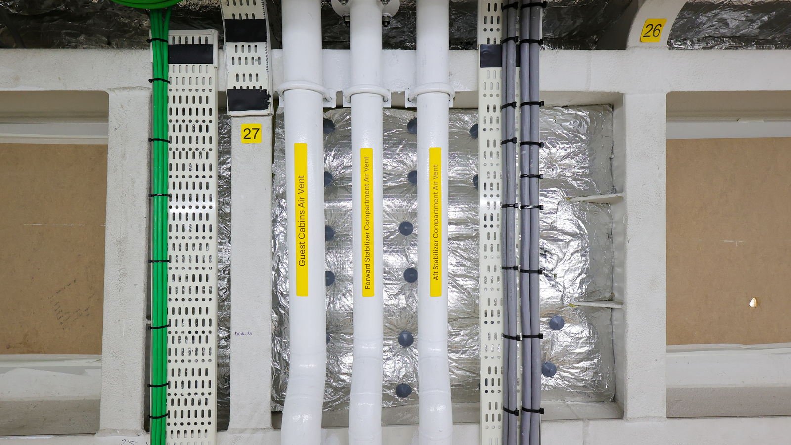 Three white vent risers running up a bulkhead, each labelled for the compartment it serves, between two cable trays and against foil-faced insulation carrying numbered frame tags