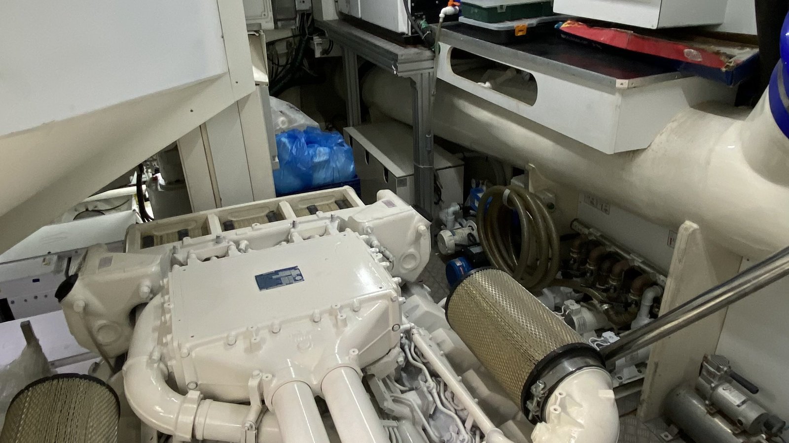 A machinery space photographed as found, the main engine and its manifold below a lagged exhaust run, with mesh air filters, a coiled hose and stores left on the deck plates