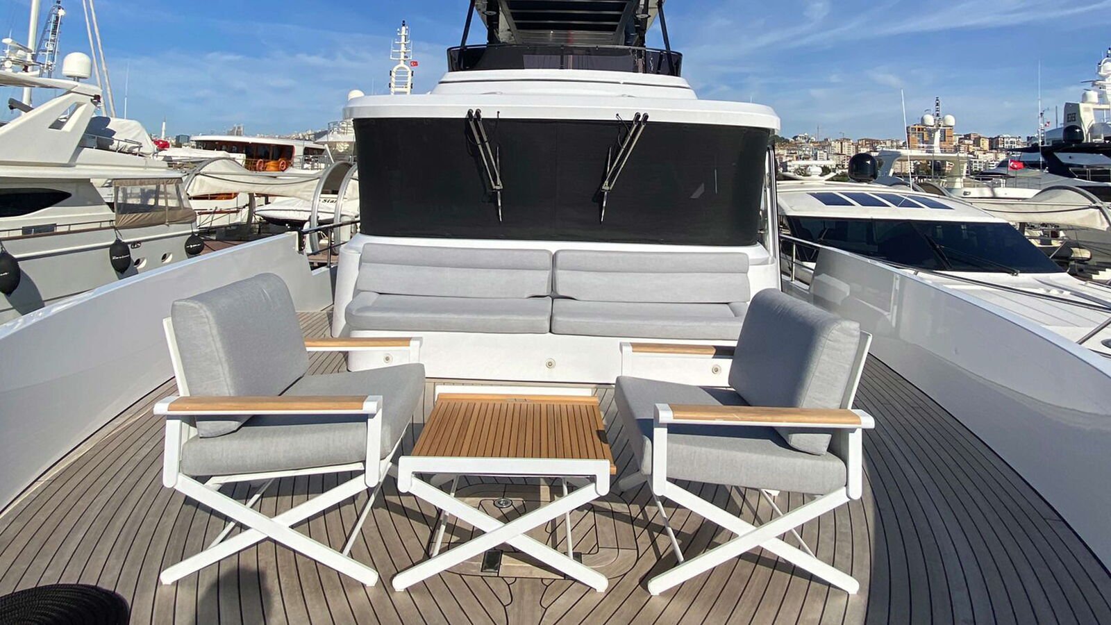 The forward seating area of a motor yacht in a marina berth, with neighbouring hulls close on both sides and a hillside city behind.