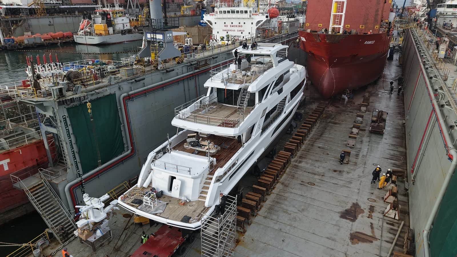 A motor yacht standing dry in a floating dock during a refit, access stairs and staging rigged along her side and the dock floor clear around her hull