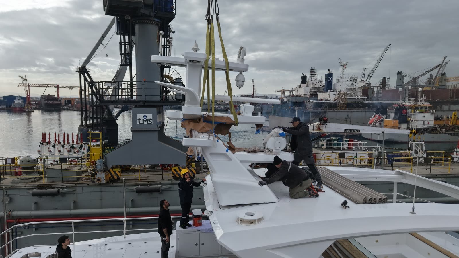A yard crane lowering a fabricated superstructure section onto a yacht flybridge while yard hands guide it into place