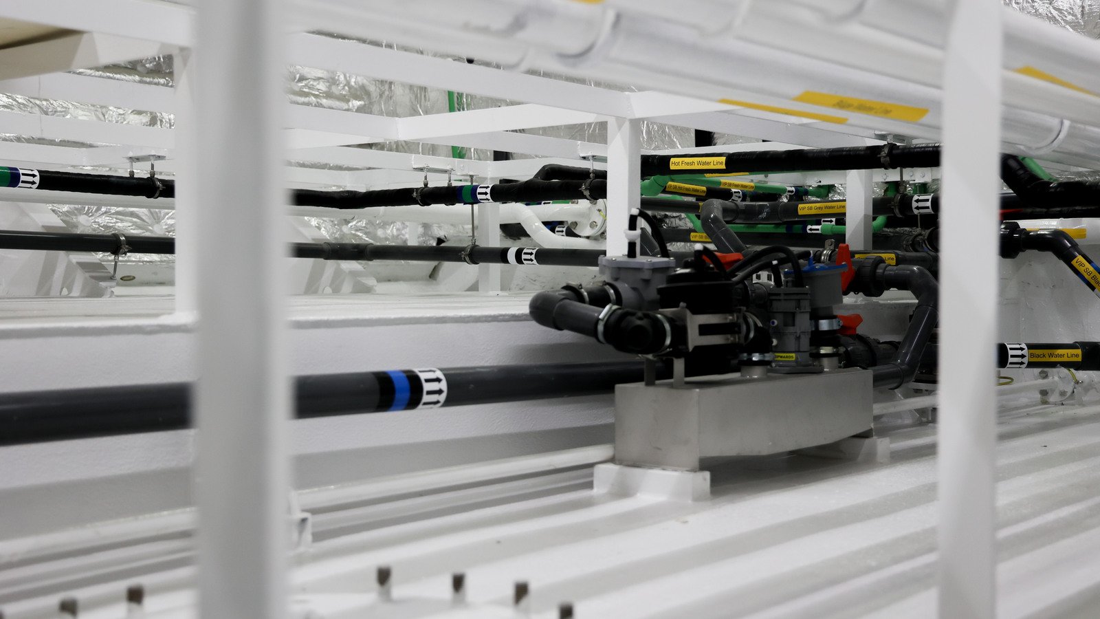 A grey pump-and-valve unit on a stainless base plate, bolted down to a low plinth in a white-painted void, photographed through the sole bearers that cross the foreground out of focus; flexible couplings, hose clamps and a red lever valve join it to the black and green lines carried on the framing behind, and a row of short studs stands proud of a plate in the blurred foreground.