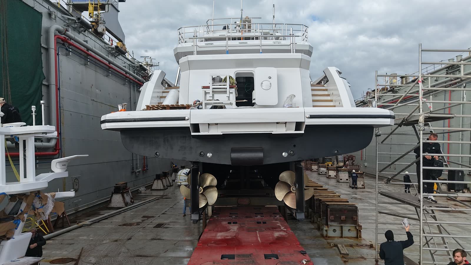 A motor yacht up on keel blocks in dry dock seen from astern, both propellers, shafts and rudders exposed, with staging rigged alongside her