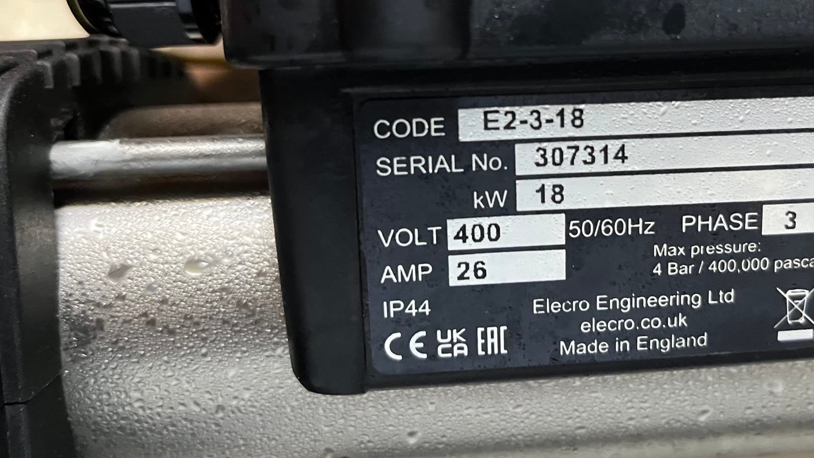 A close, slightly angled view of a manufacturer's rating plate on the end cap of a piece of installed plant. The plate is black with white boxed fields carrying a code, a serial number, a rating in kilowatts, a voltage with a fifty and sixty hertz note, a phase figure, a current in amps and an ingress rating, with a maximum pressure given in both bar and pascals. Below them sit the maker's name and web address, the country of manufacture, and CE, UKCA and EAC marks beside a crossed-out wheeled bin. A brushed stainless body beaded with condensation runs across the lower third of the frame, and the dark moulded casing the plate is fixed to fills the upper left.